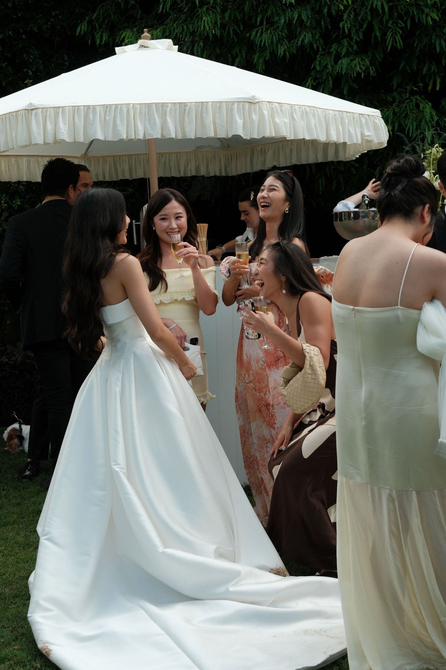 A group of wedding guests in formal attire gather under a white umbrella during an outdoor celebration.