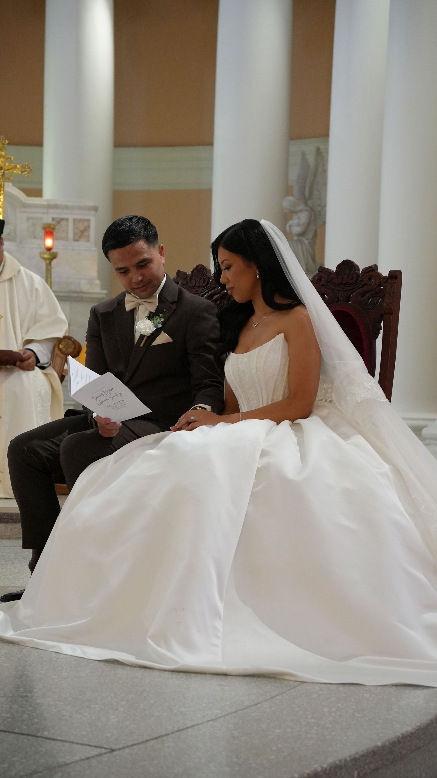 Bride in white gown and groom in brown suit sit together during church wedding ceremony, reading from a paper.