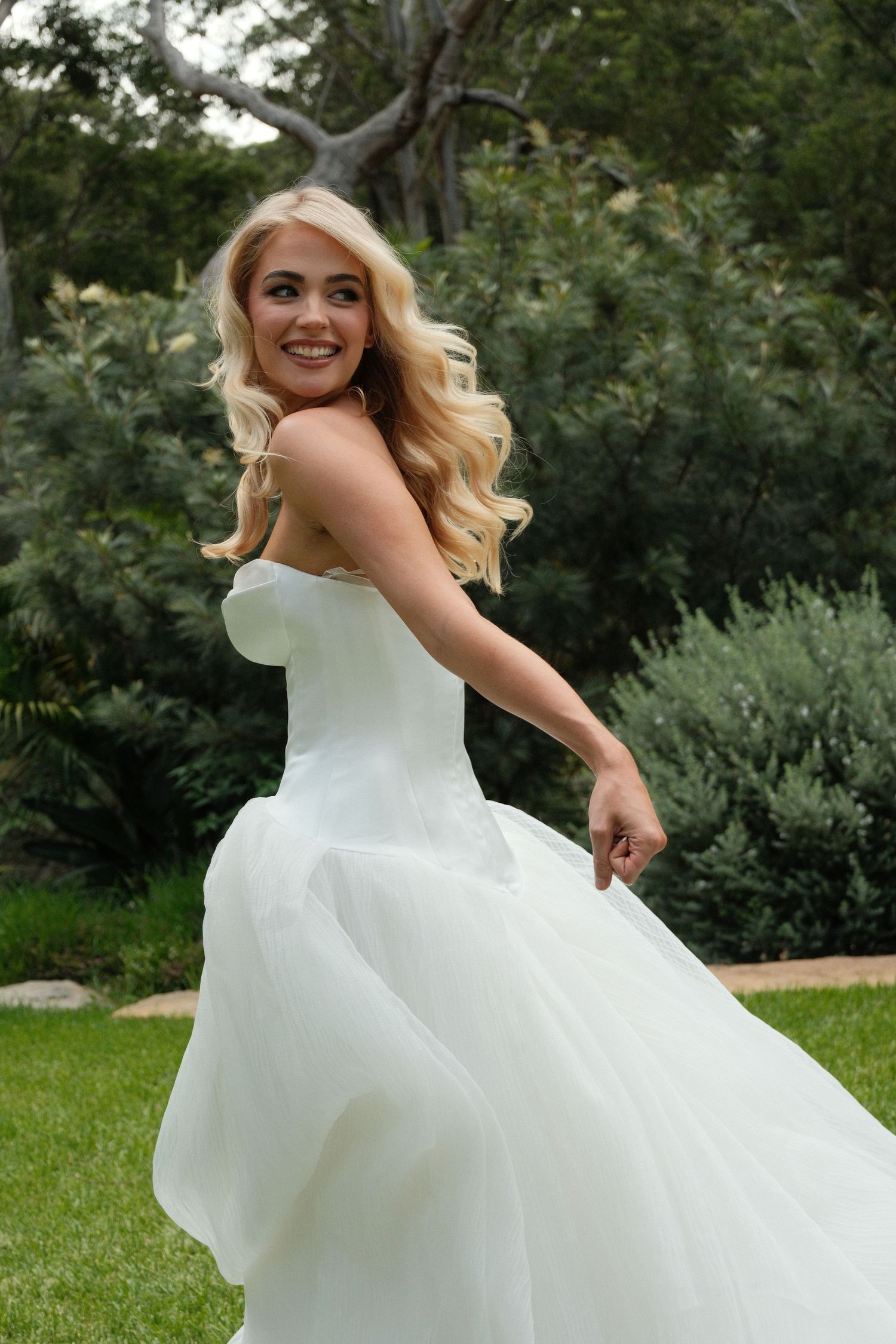 A bride in a flowing white ballgown twirls playfully in a lush garden setting, making her dress billow in the breeze.