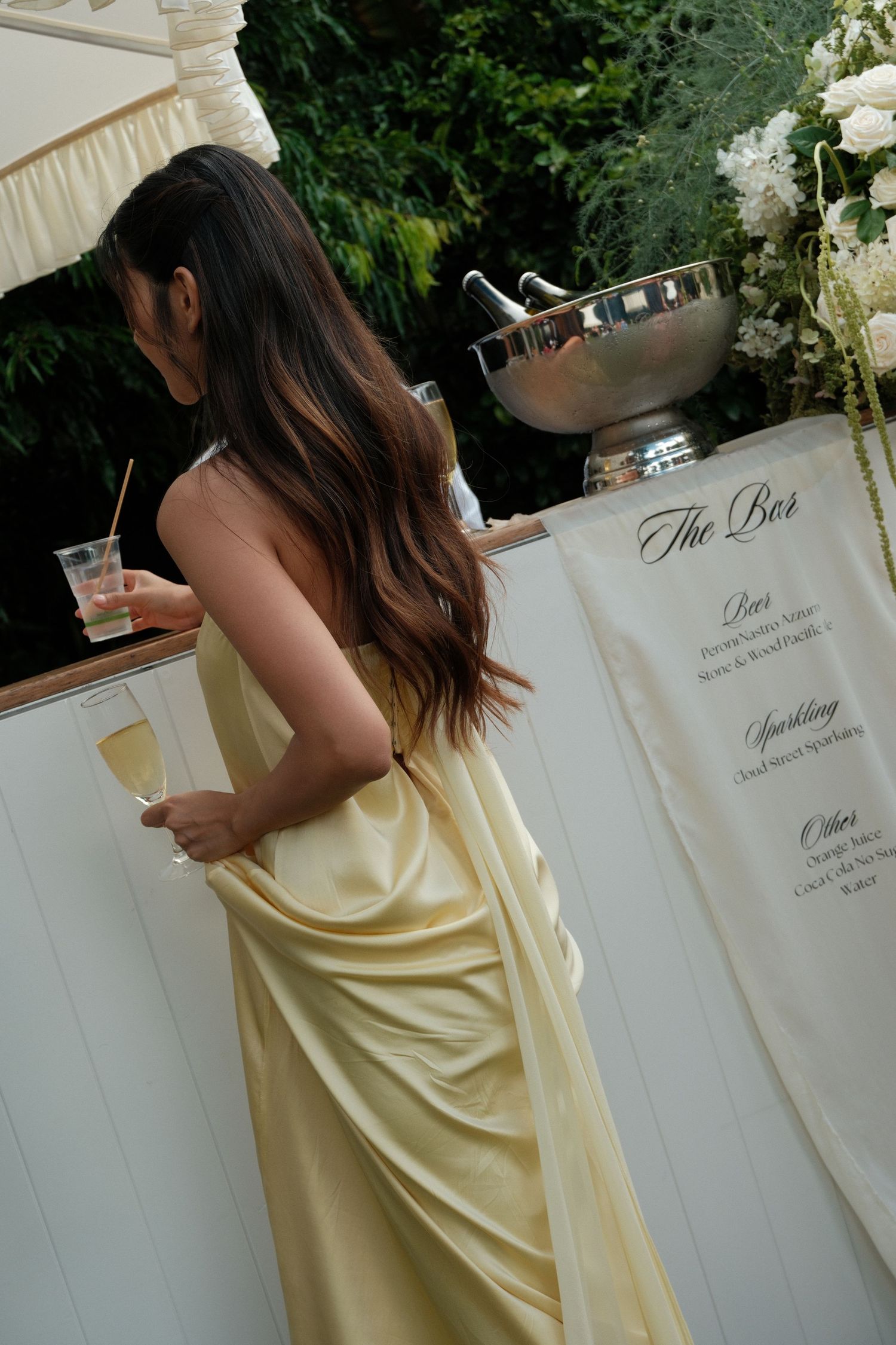 Person in flowing yellow gown stands at outdoor bar with cocktail during elegant garden party.