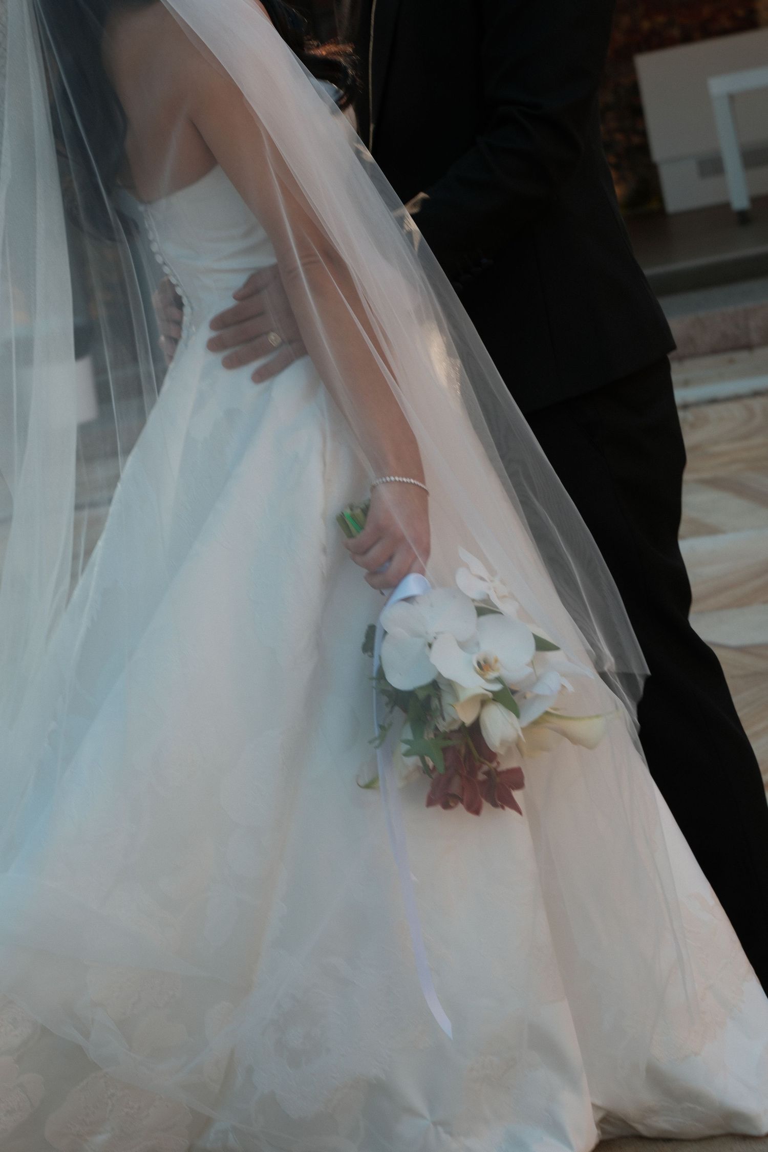 Bride in white strapless gown holding white orchid bouquet, groom in black suit, wedding ceremony.