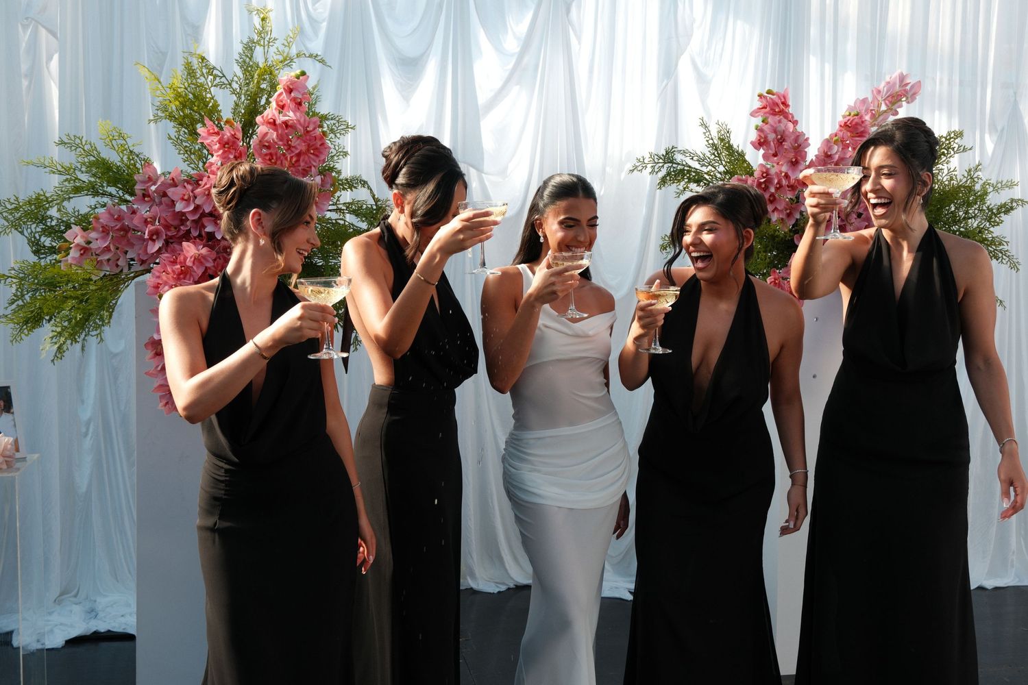 Bridesmaids in black dresses celebrate with the bride in white at an elegant floral wedding reception.