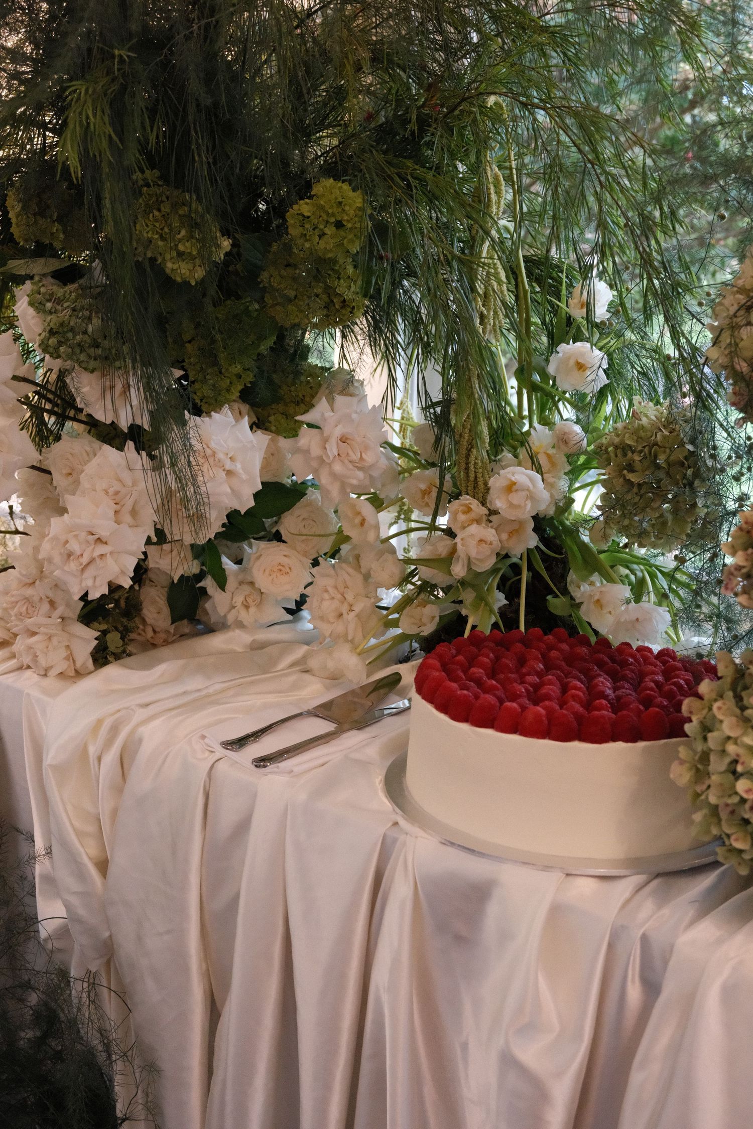 An elegant white wedding cake topped with red roses sits on a draped table with floral arrangements and greenery decorations.