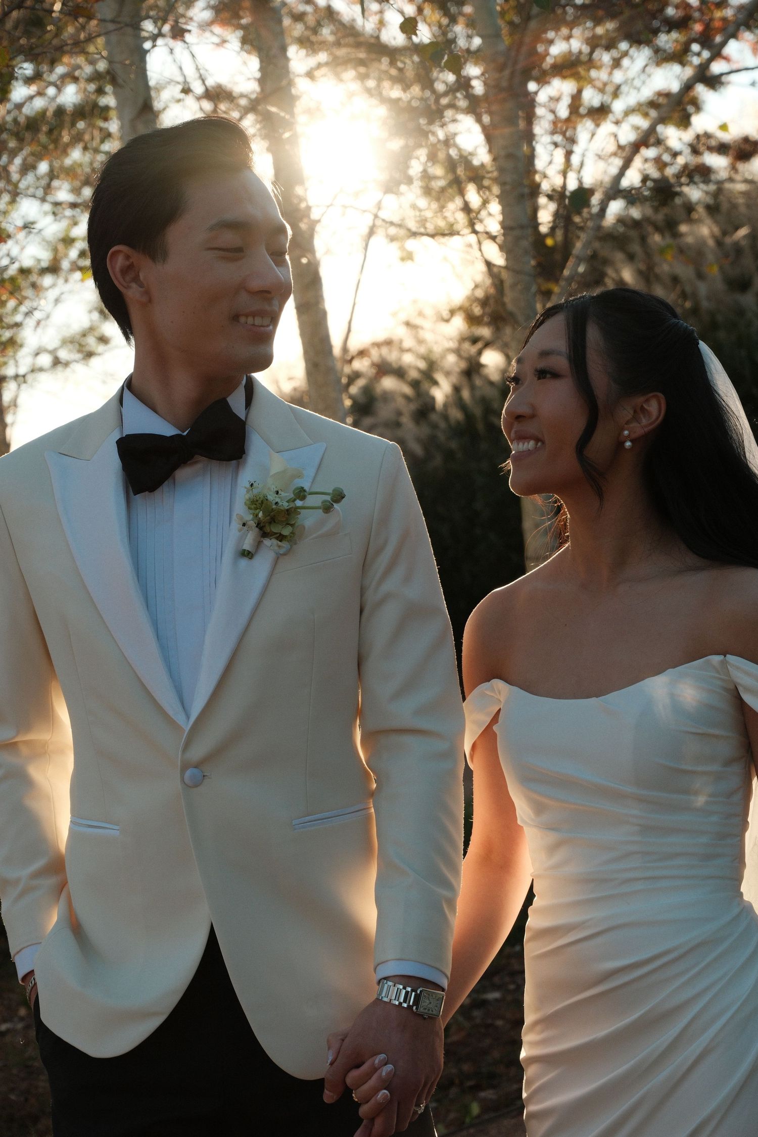 Smiling bride and groom holding hands in golden sunlight, groom in white tuxedo, bride in strapless wedding gown.