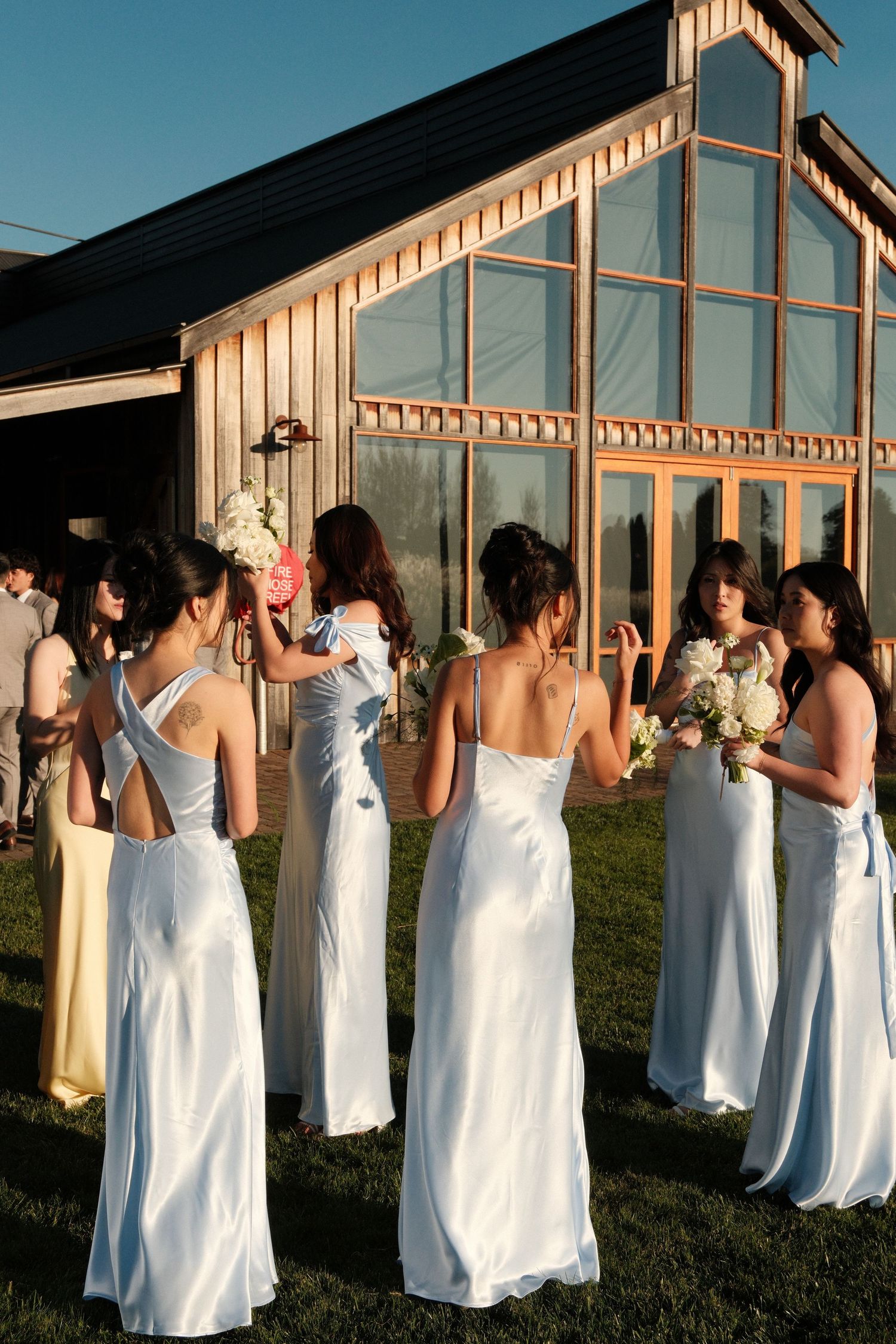 Bridesmaids in light blue satin gowns holding bouquets stand outside a rustic barn wedding venue at golden hour.