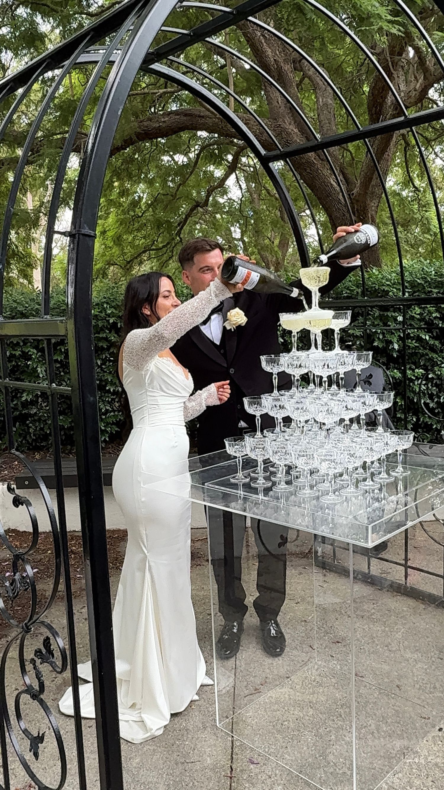 Wedding party pouring champagne into a tower of crystal glasses under an outdoor gazebo.
