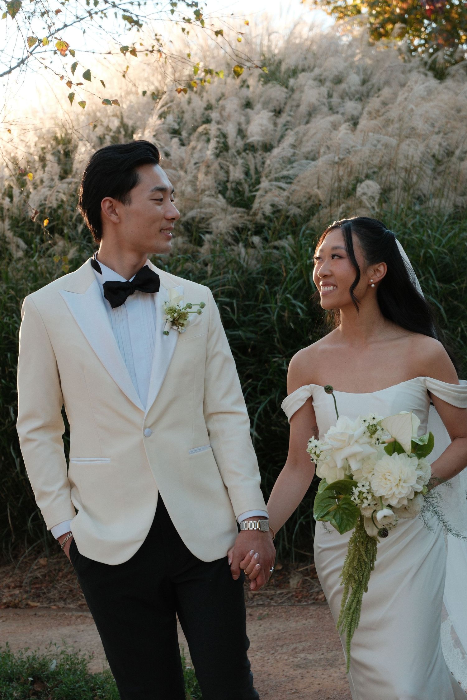 Smiling bride and groom in cream tuxedo hold hands in front of tall ornamental grasses at sunset wedding.
