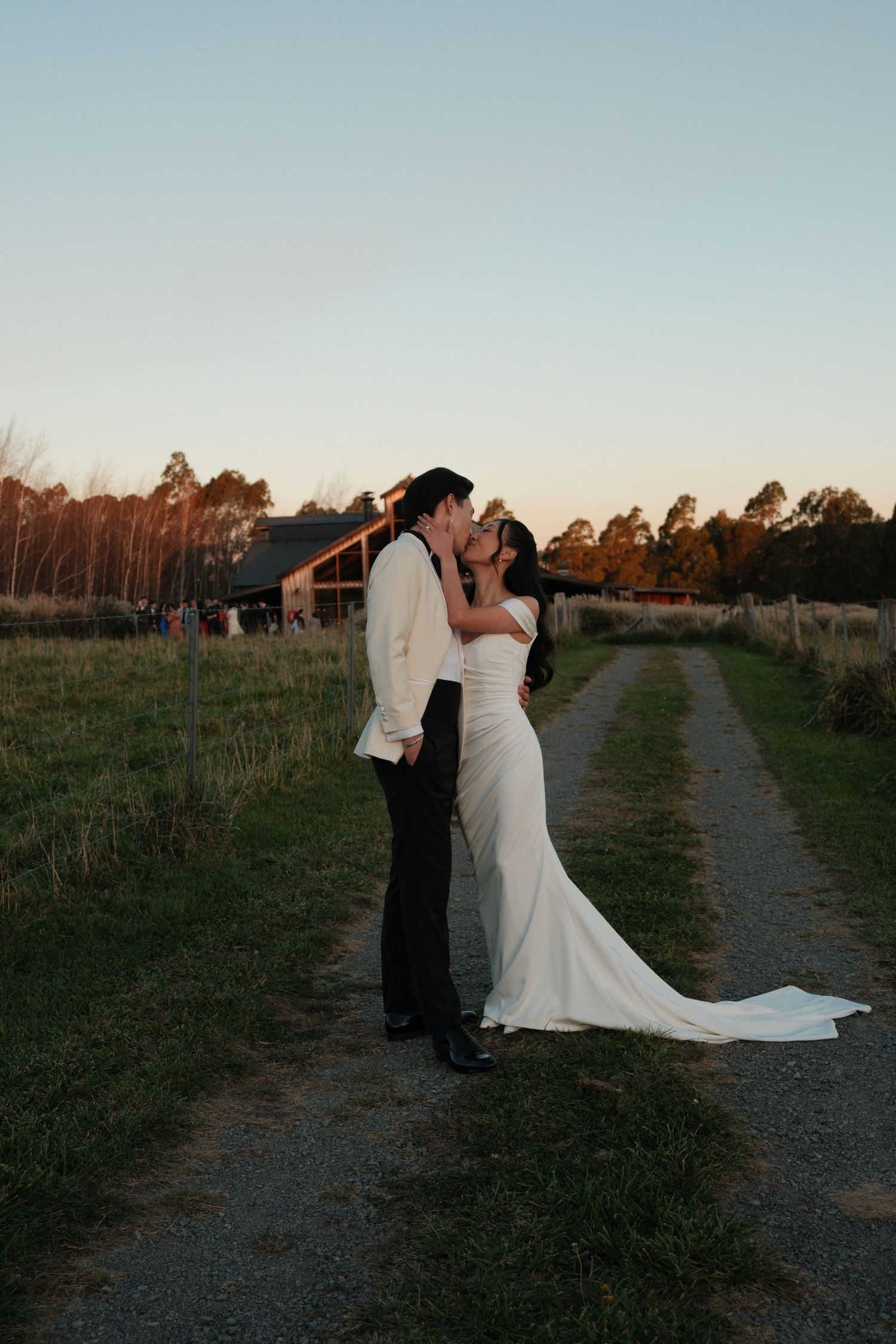 Bride and groom sharing a kiss at sunset on a rural gravel path, with a barn and trees in the background.