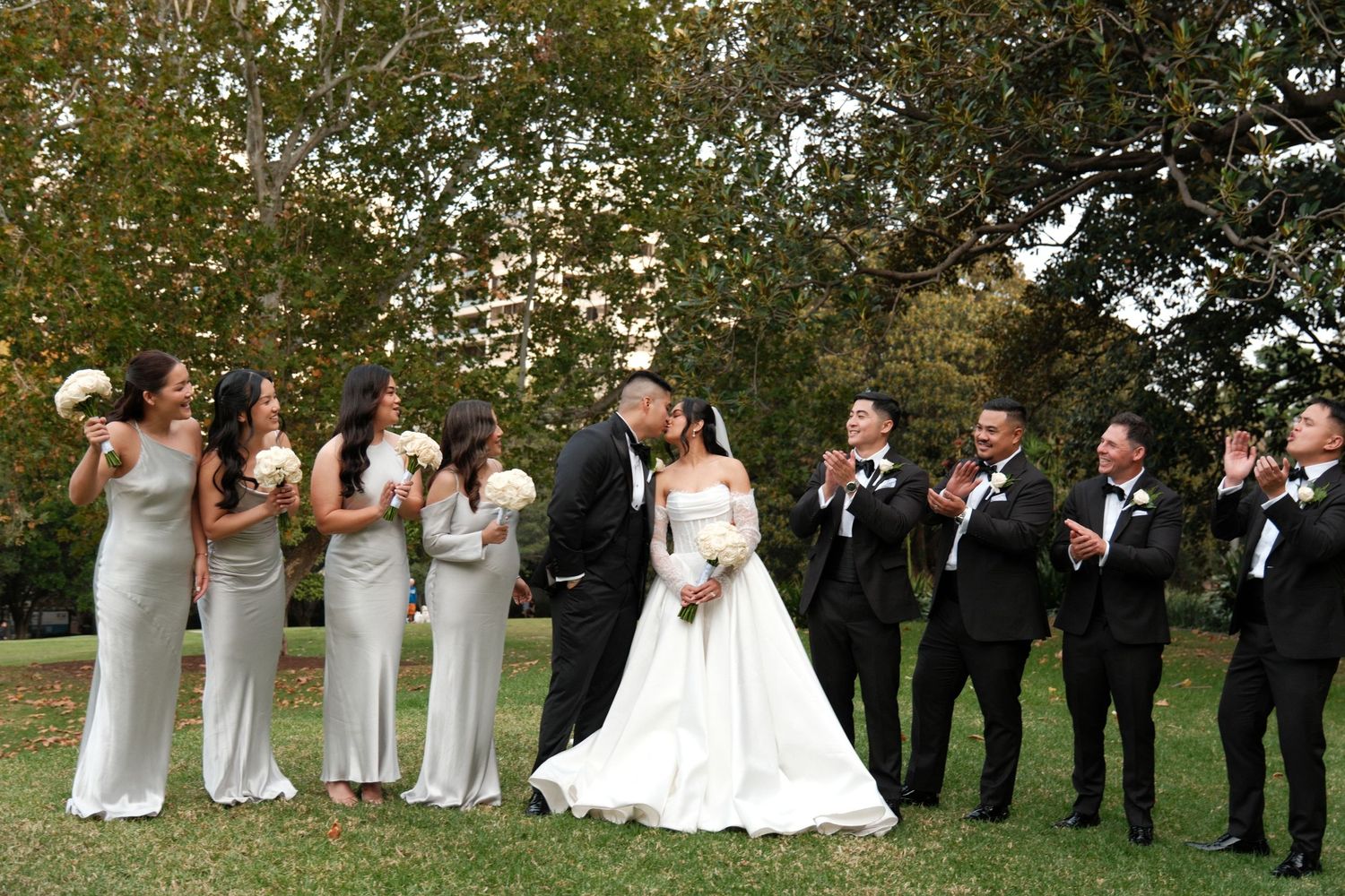 Wedding party poses outdoors, bride and groom kissing center, bridesmaids in silver gowns and groomsmen in black tuxedos.