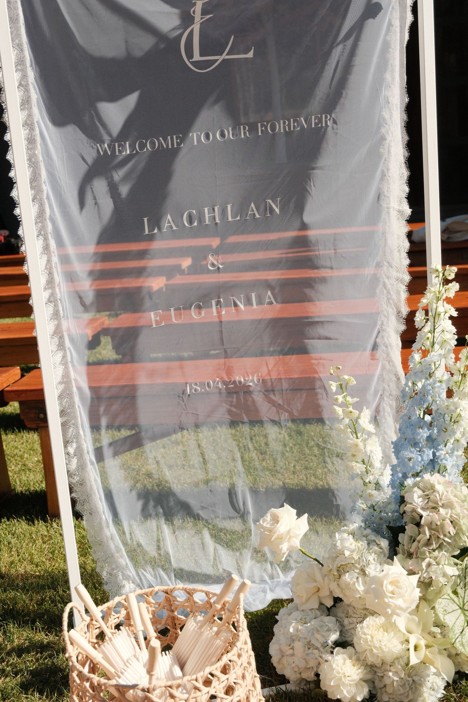 Elegant outdoor wedding welcome sign for Lachlan and Eugenia on acrylic with white floral arrangements and wicker basket.
