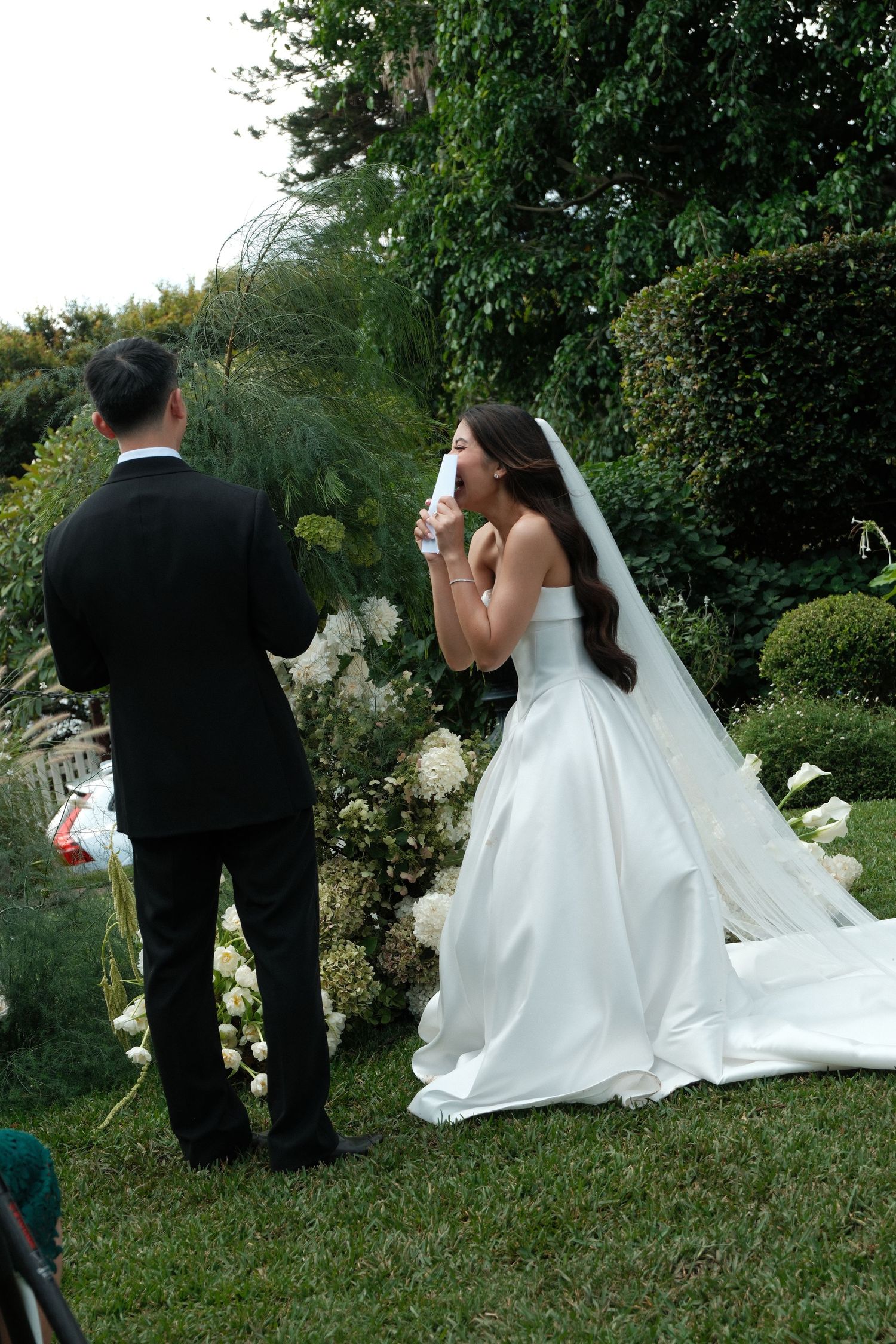 A wedding couple poses in a lush garden setting with flowing white dress and veil.
