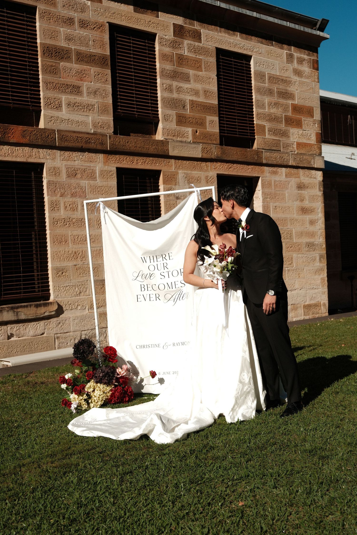 Bride and groom kissing outdoors beside a decorative wedding banner on green grass near a stone building.