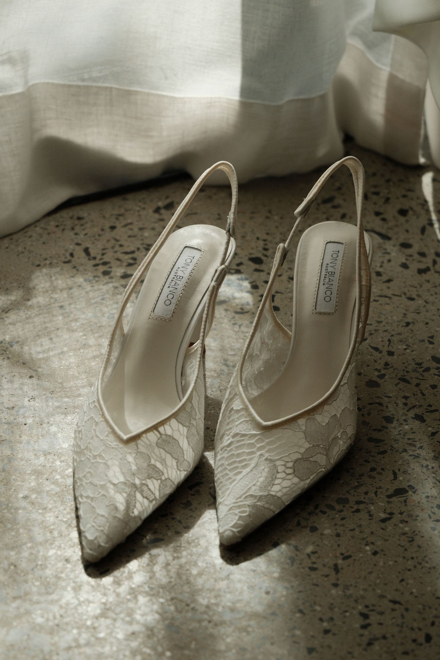Elegant ivory lace Jimmy Choo slingback heels on marble floor with sheer white fabric backdrop.