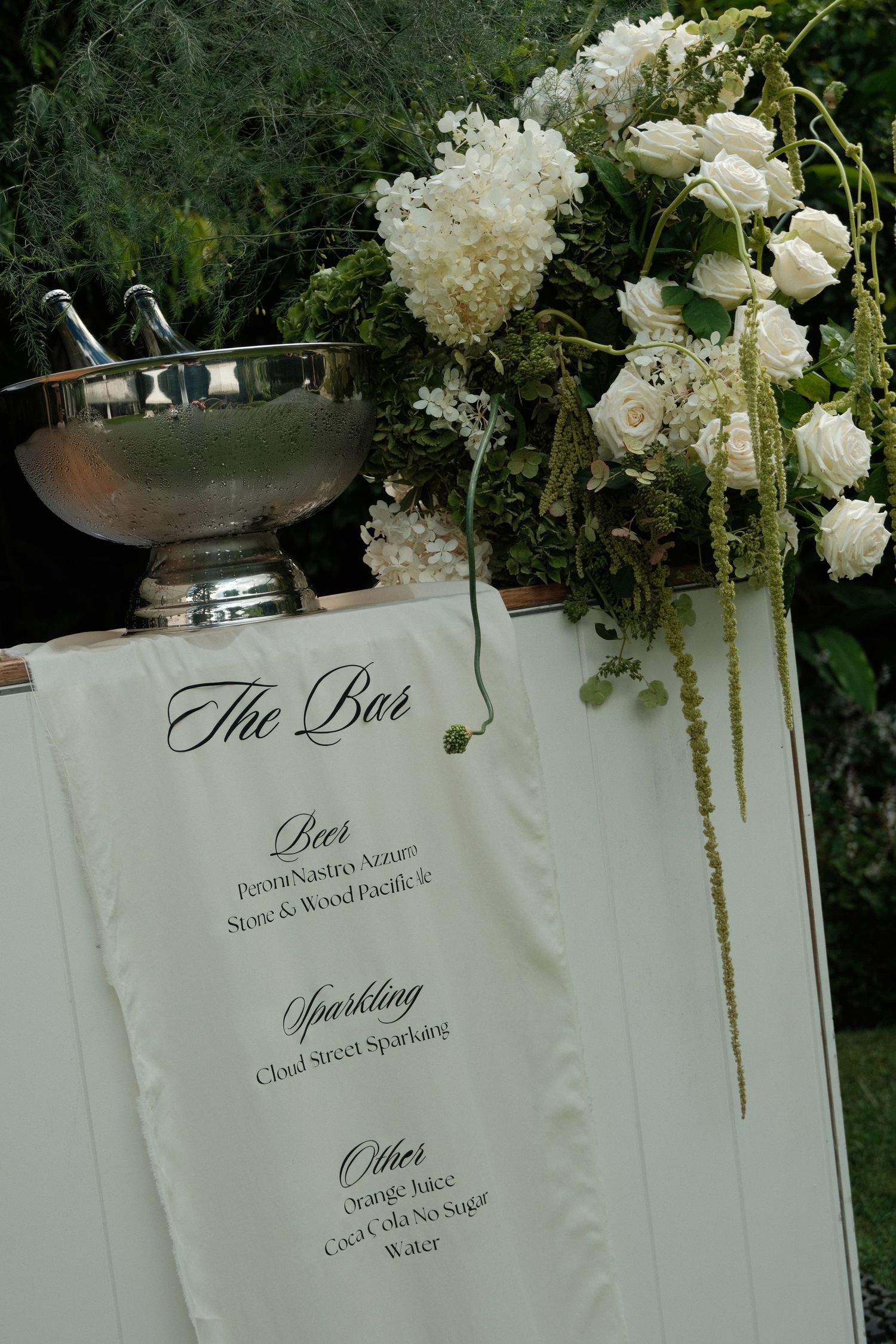 Elegant white floral arrangements and wedding menu cards displayed against a dark green garden backdrop.