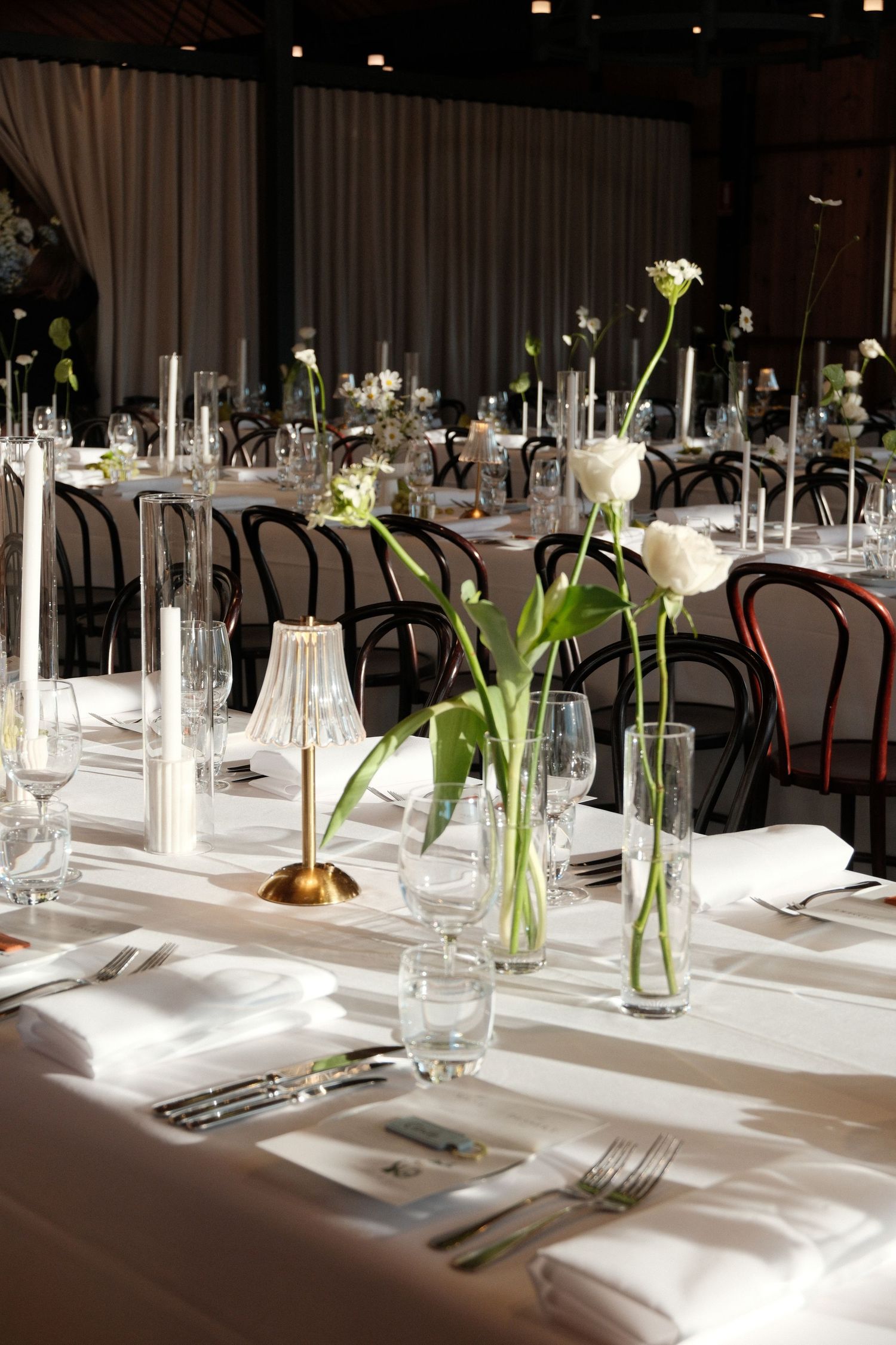 Elegant wedding reception tables with white linens, tall glass vases holding white tulips, candles, and black bentwood chairs.