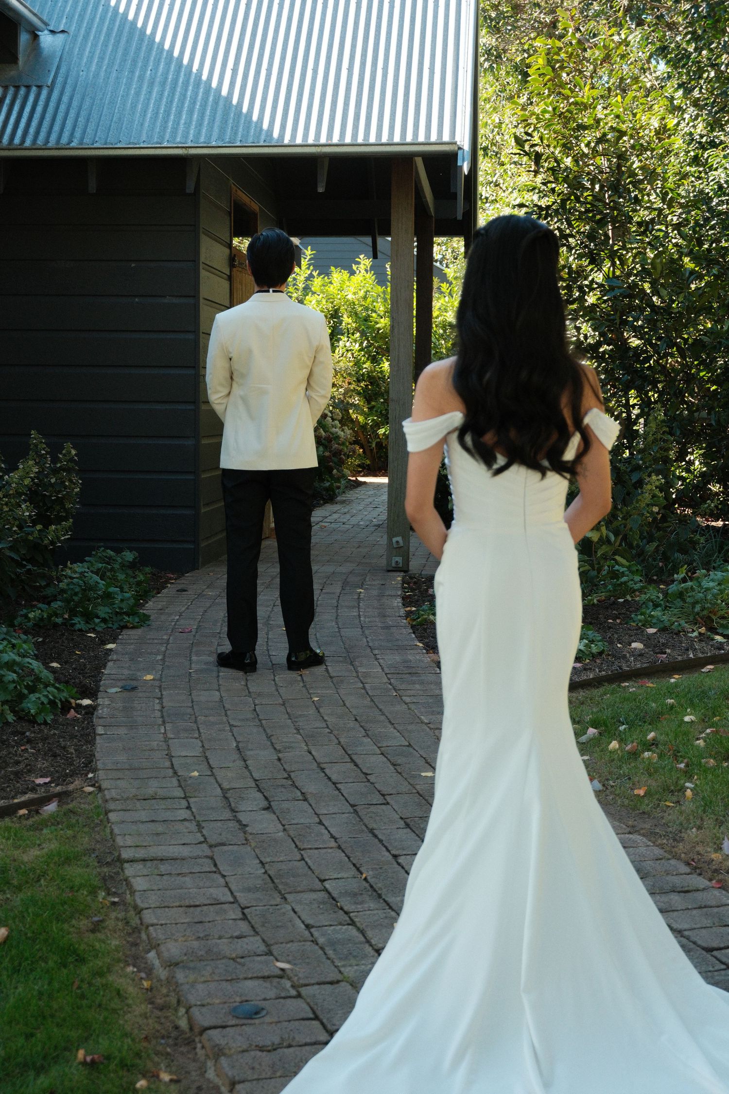 Bride in white off-shoulder gown and groom in ivory suit stand apart on brick path during wedding first look.
