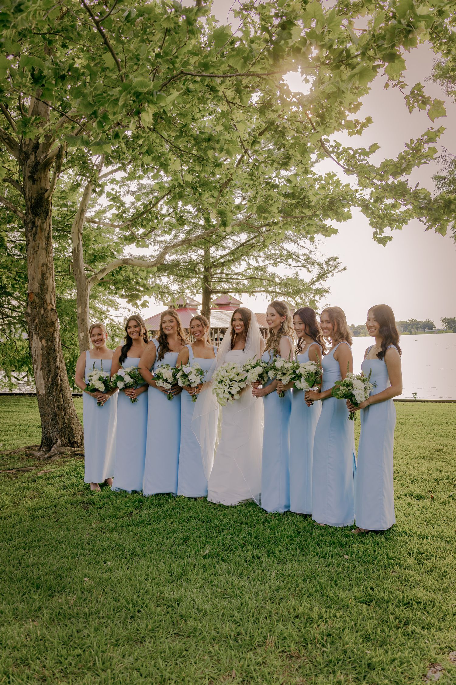 A row of bridesmaids in light blue dresses stand together on a grassy lawn under a large tree.