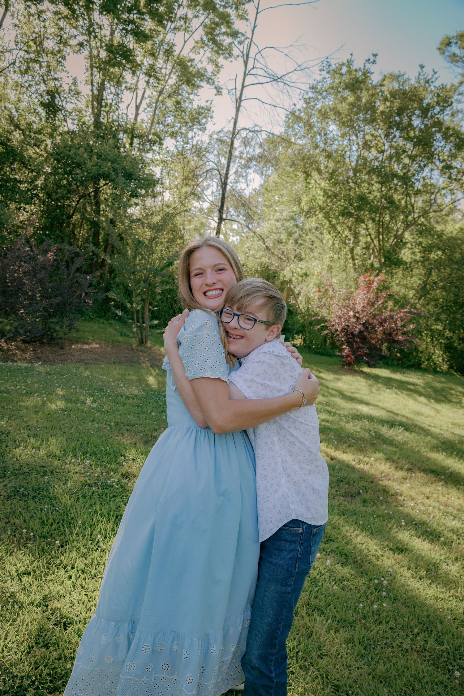 A pair wearing light colored clothing embrace while laughing in a grassy outdoor setting.