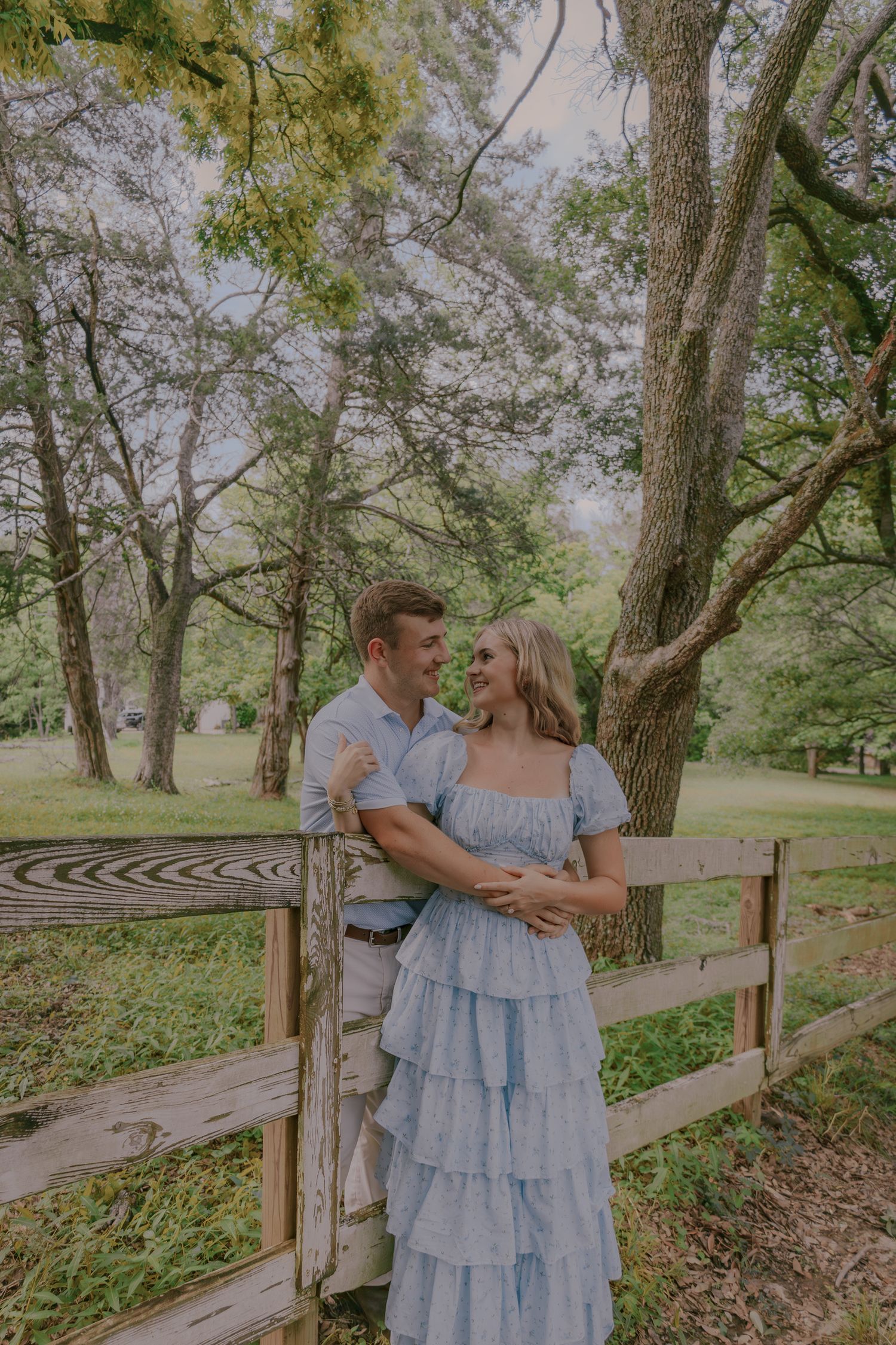 A couple shares tender moments along a wooden fence path surrounded by lush trees.