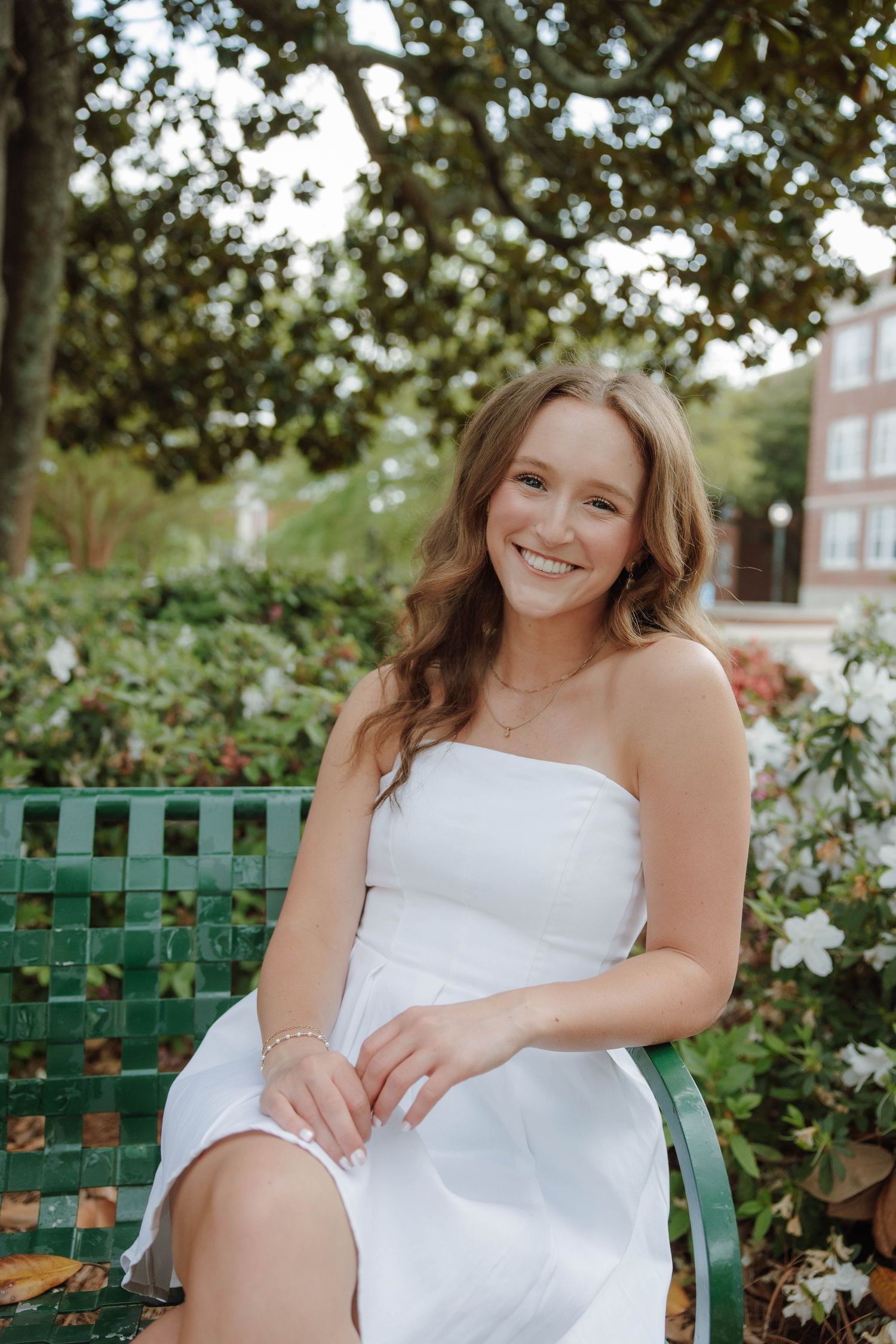 Young woman in white strapless dress smiling on green bench surrounded by lush garden greenery and flowers.