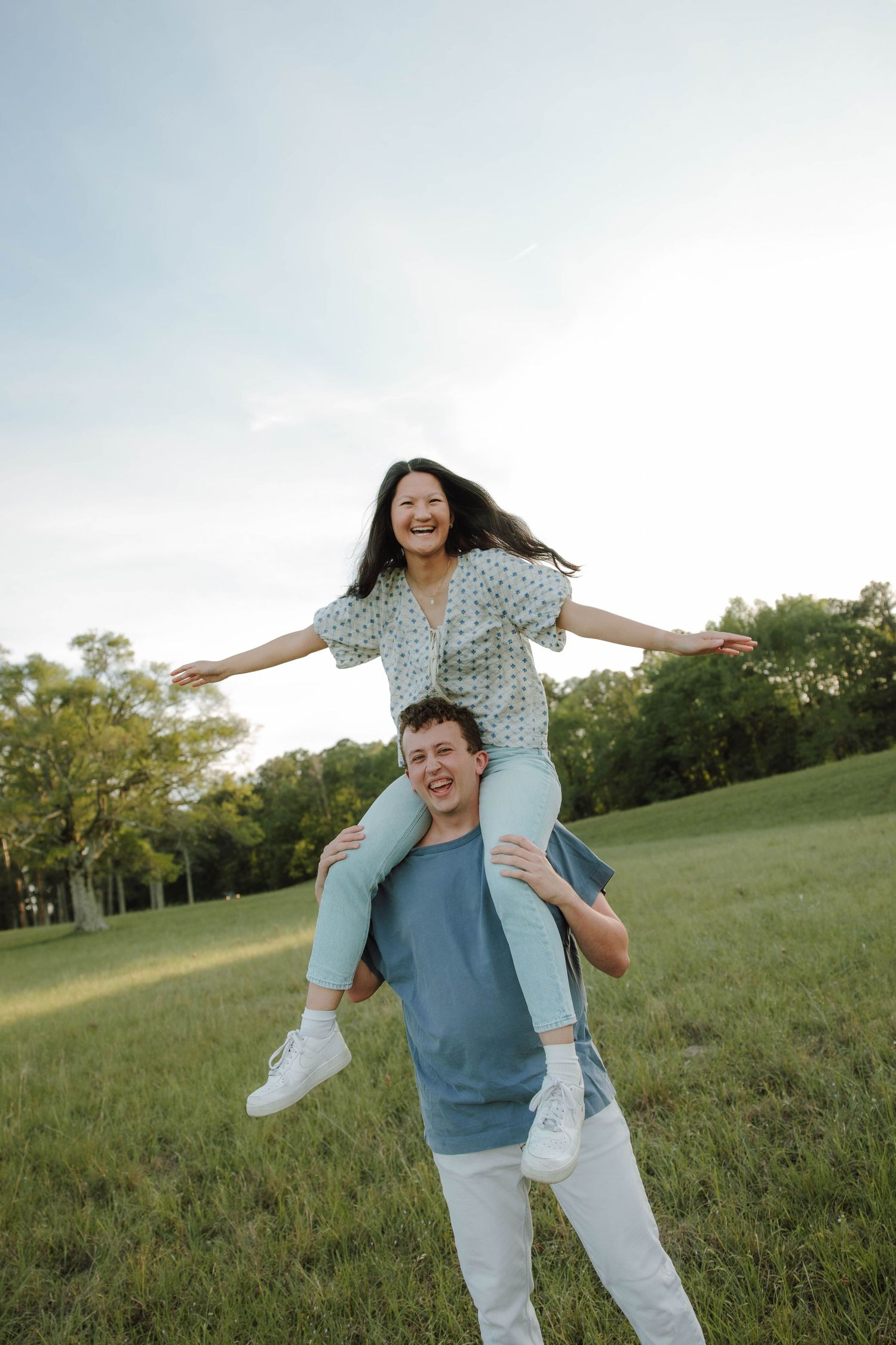 A joyful woman sits on a man's shoulders with arms outstretched in a green park field under a cloudy sky.