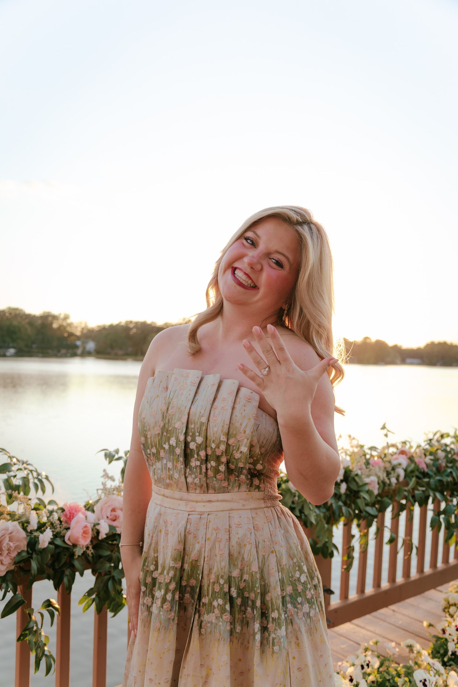 Person in a floral dress twirling on a wooden deck overlooking a lake at sunset.