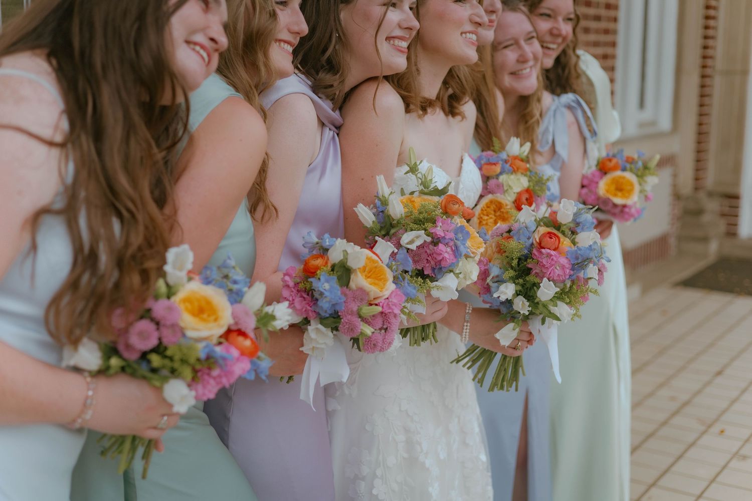 Bride and bridesmaids in pastel dresses holding colorful spring bouquets with pink, yellow, and blue flowers.