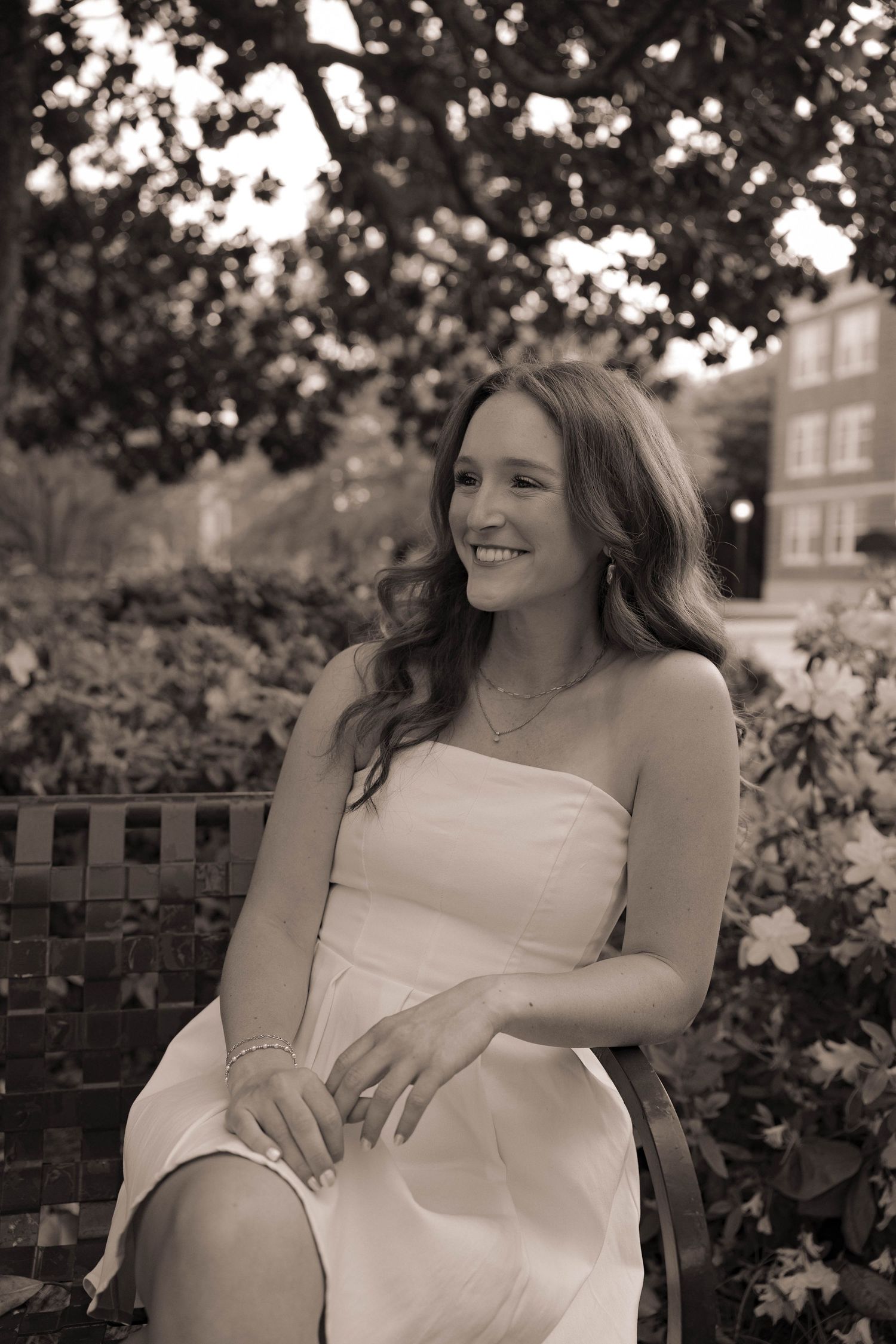 Young woman in white strapless dress smiling while seated on bench surrounded by lush garden greenery, sepia tone.