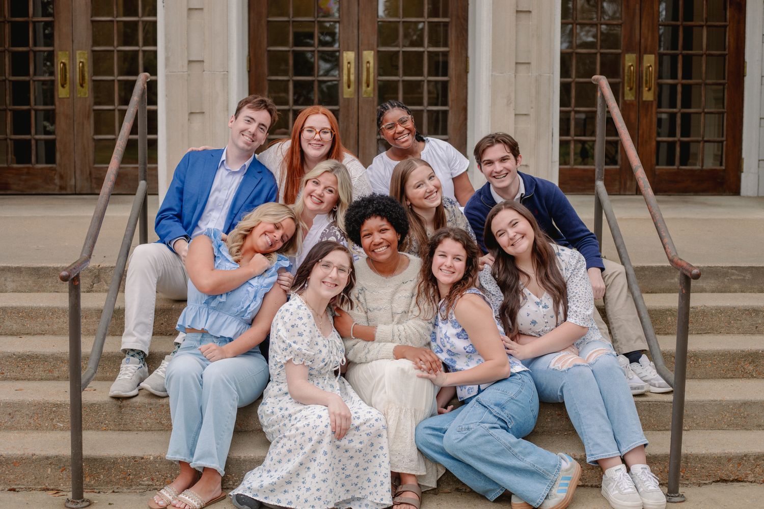A group of friends sitting together on stone steps outside a building with large windows.