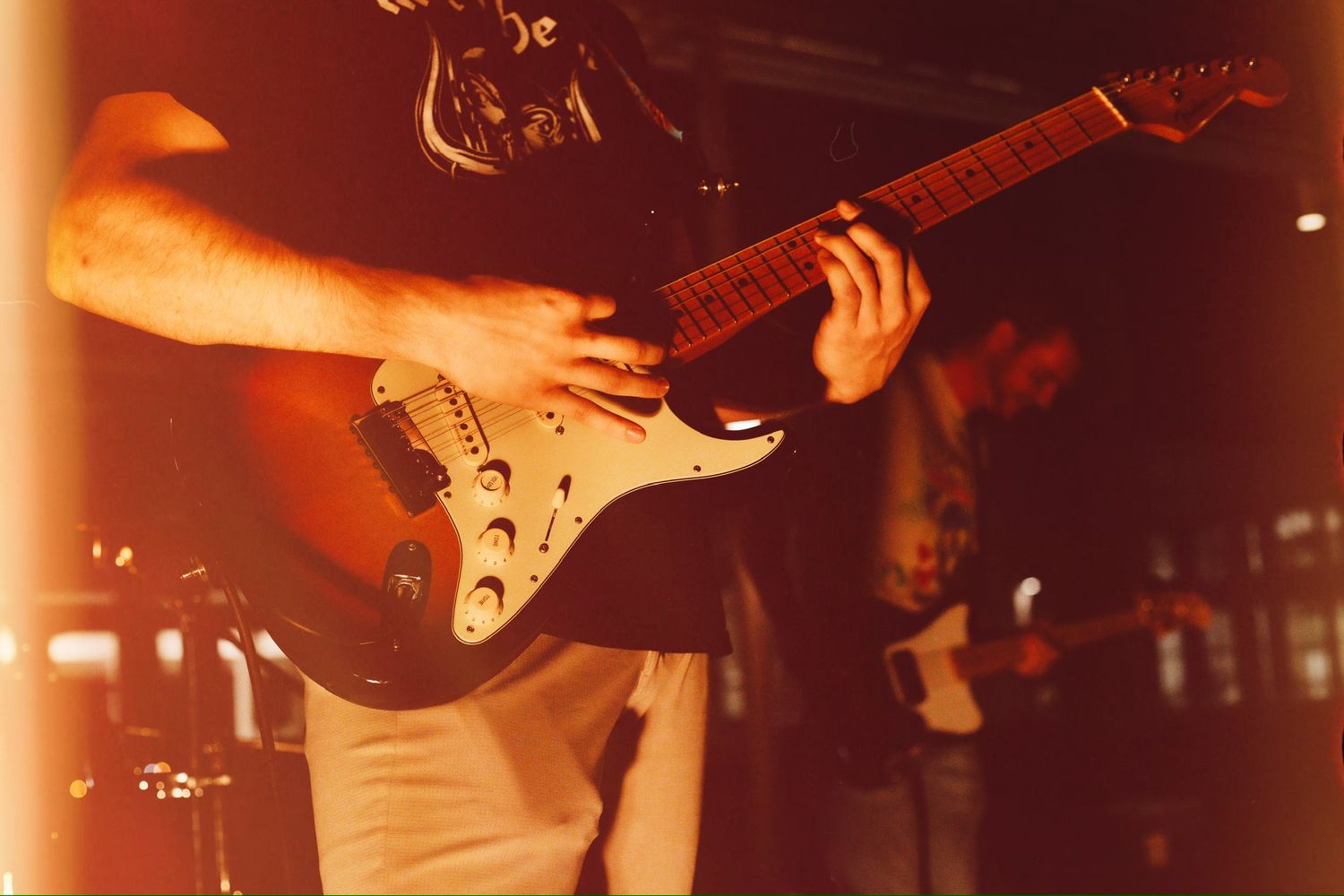 Electric guitarist playing a Stratocaster guitar in warm orange stage lighting.