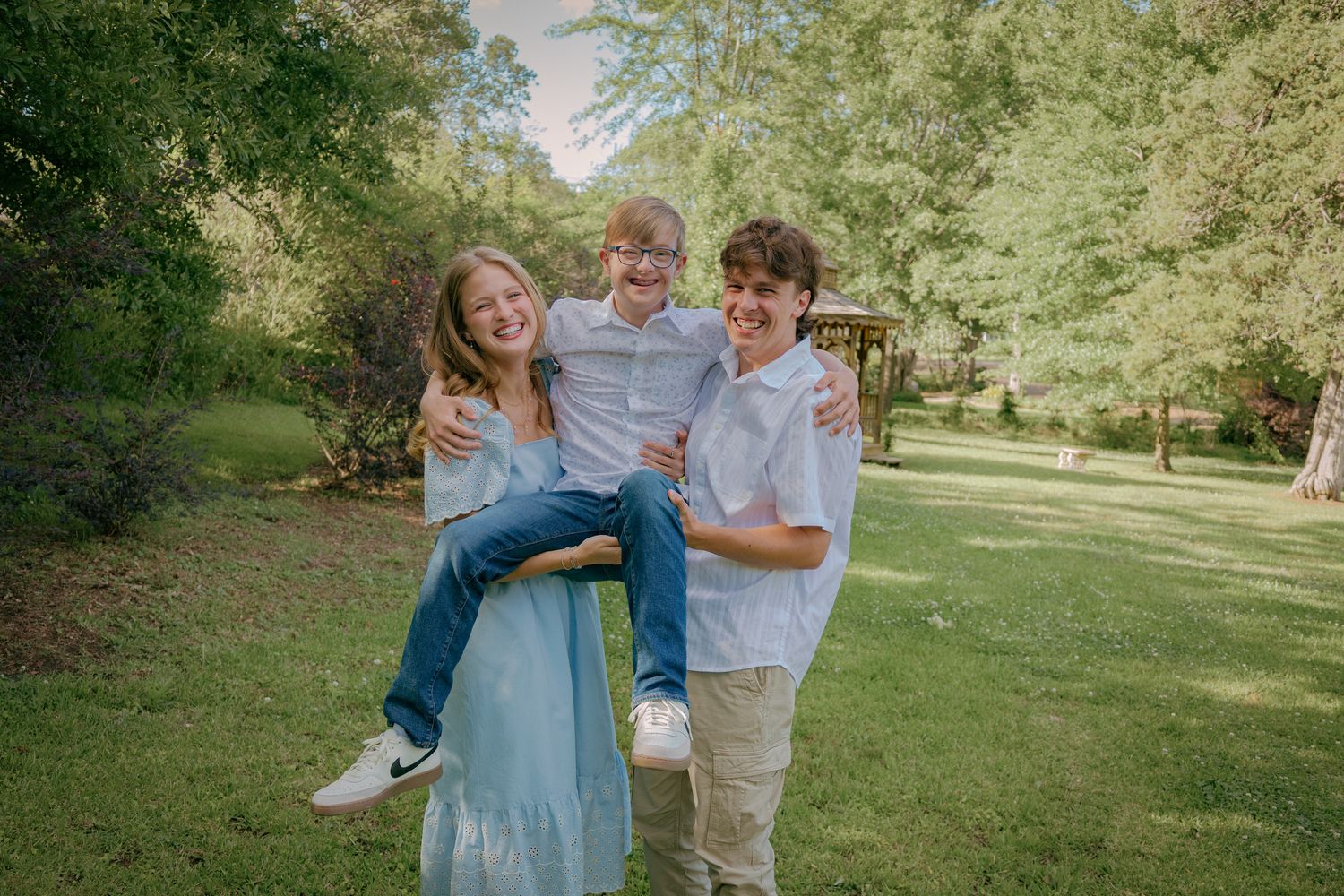 A group in casual attire poses playfully in a sunny outdoor park setting with trees in the background.