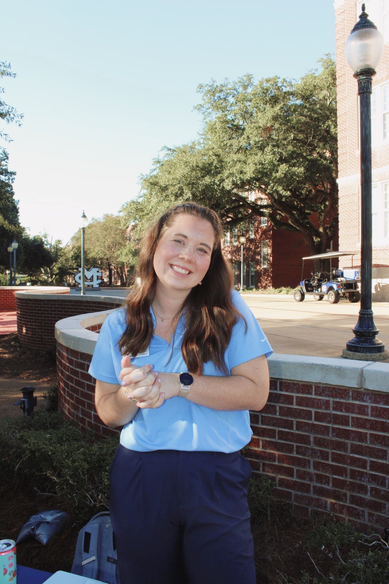 Person in light blue uniform top and navy bottoms poses cheerfully on a brick wall.