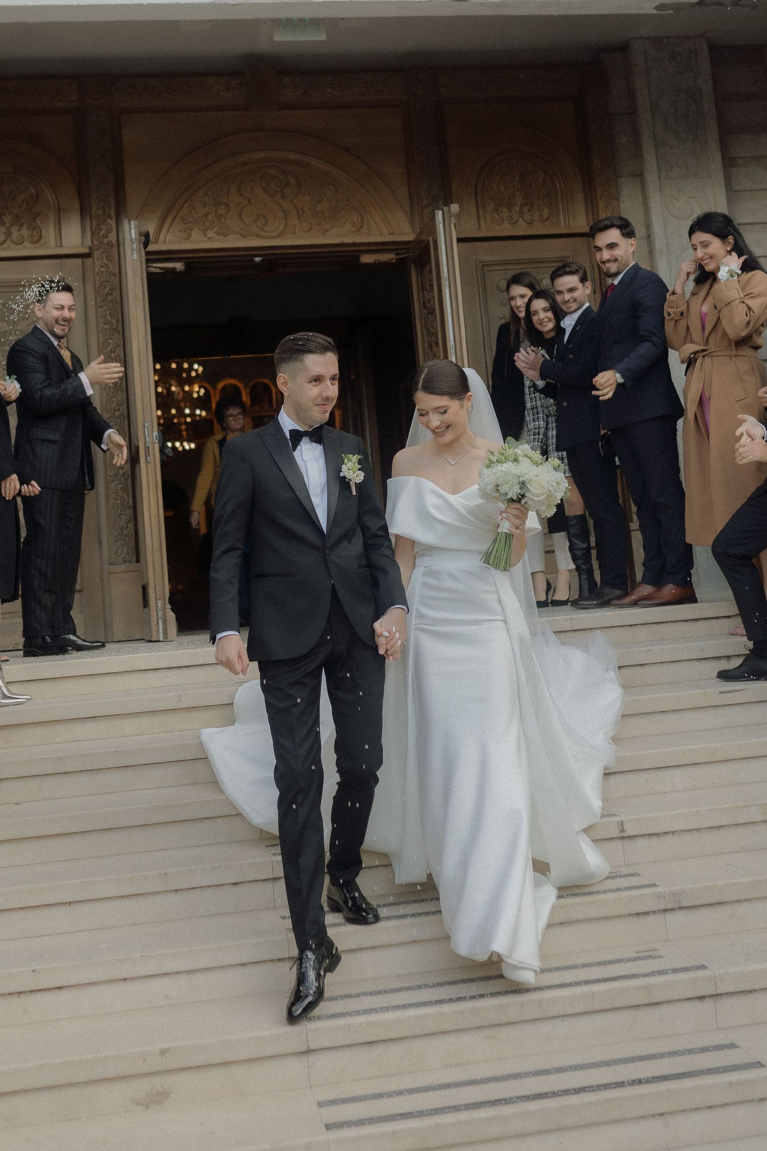 A wedding couple descends stone steps outside a building while guests watch from the doorway.