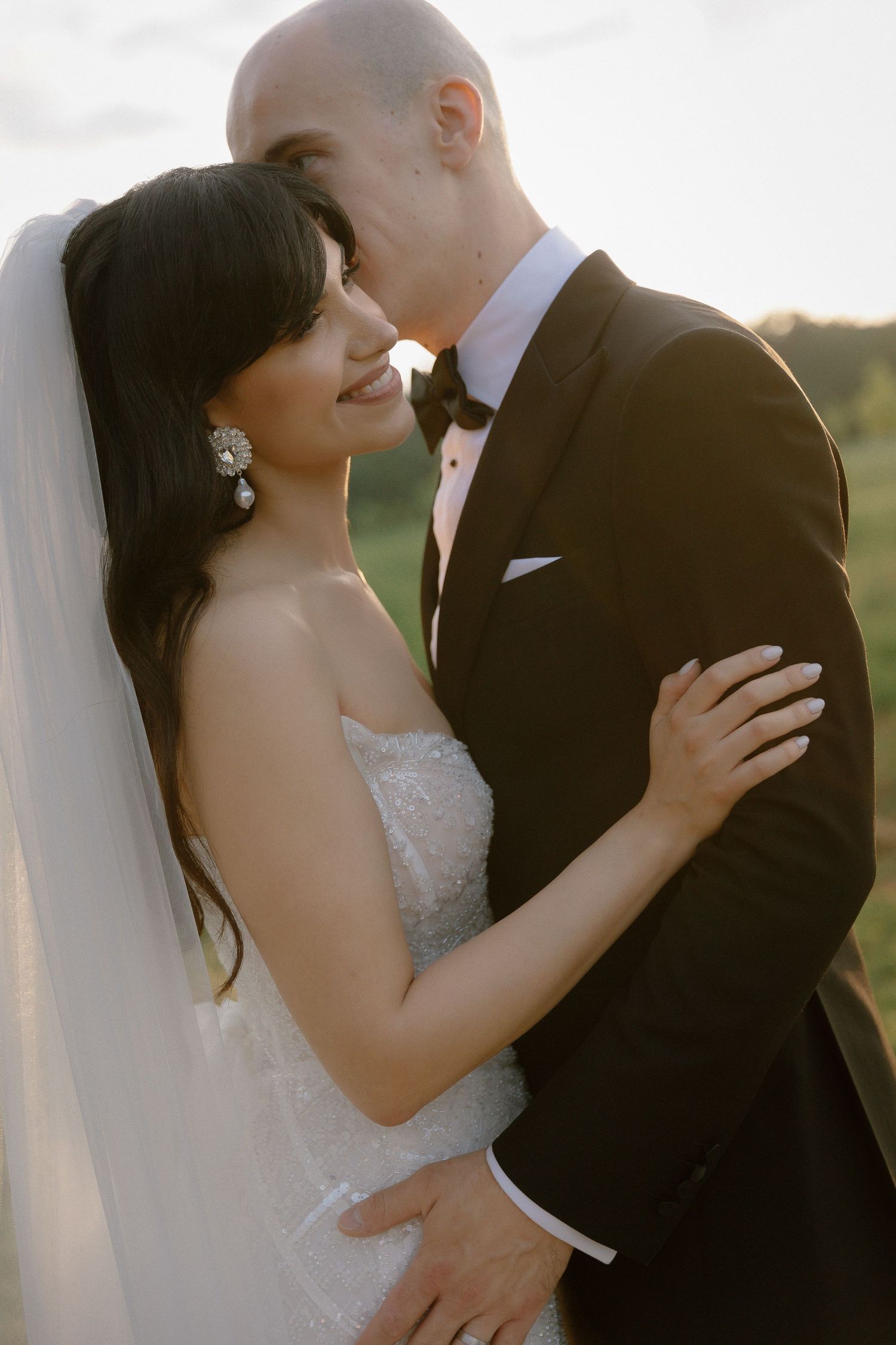 A couple in wedding attire embraces intimately during their sunset photoshoot, with the bride's veil flowing in the breeze.