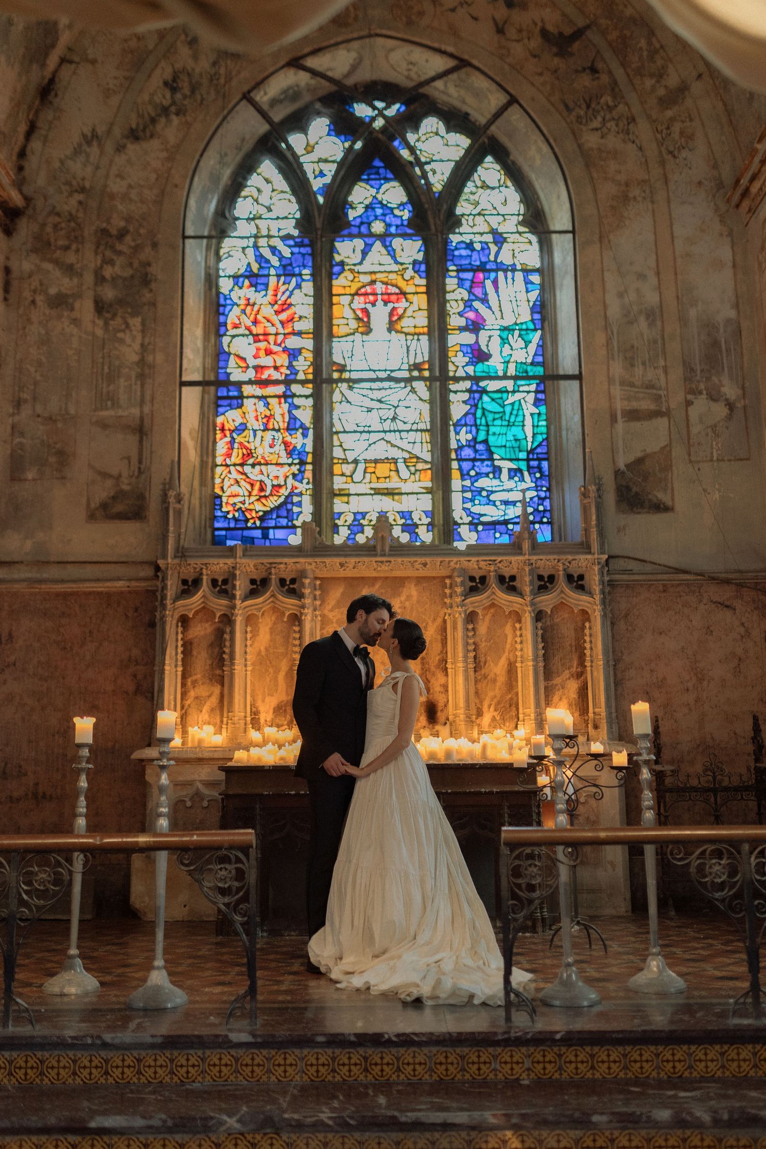 A romantic wedding couple stands before stunning stained glass windows in a historic stone chapel with glowing candles.