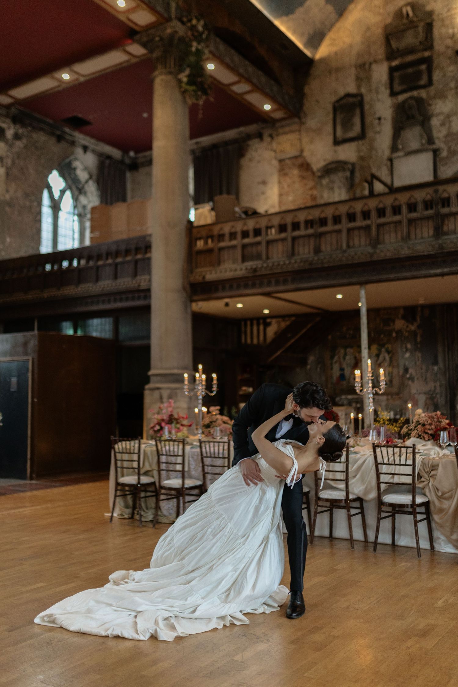 A couple shares a romantic dance in an elegant historic venue with stone columns and candlelit reception tables.