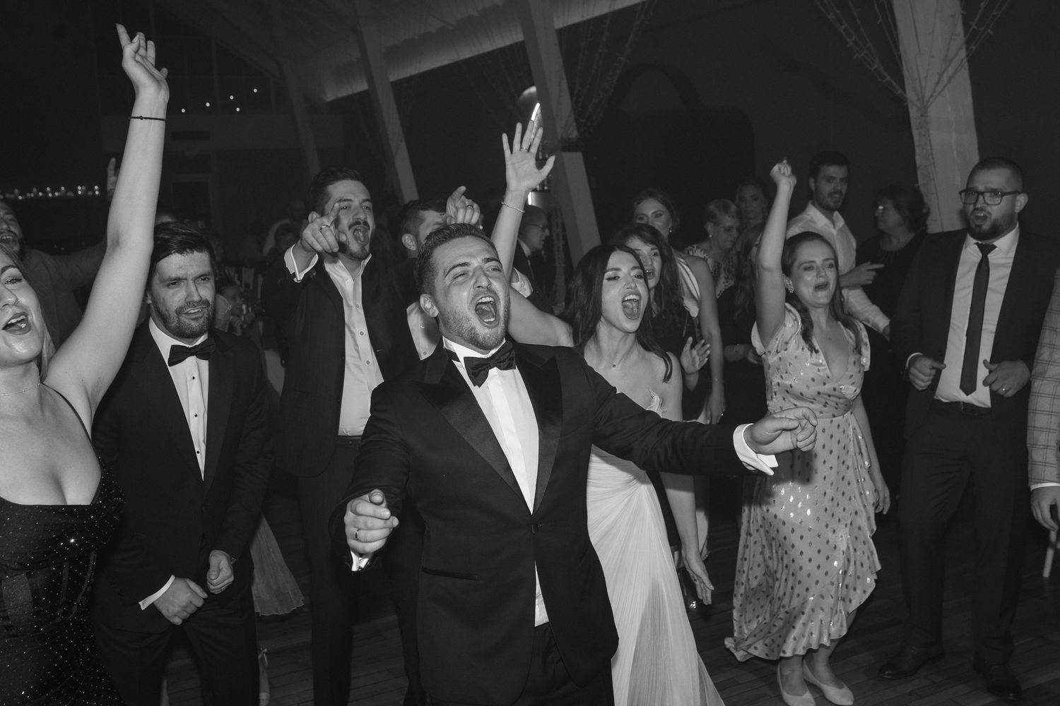 A crowd of wedding guests dancing and celebrating with their hands raised on a dark dance floor at a reception.