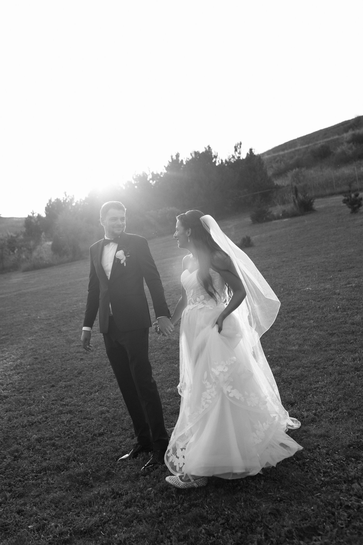 A couple in wedding attire walks together in a grassy field at sunset, their silhouettes creating a romantic scene.