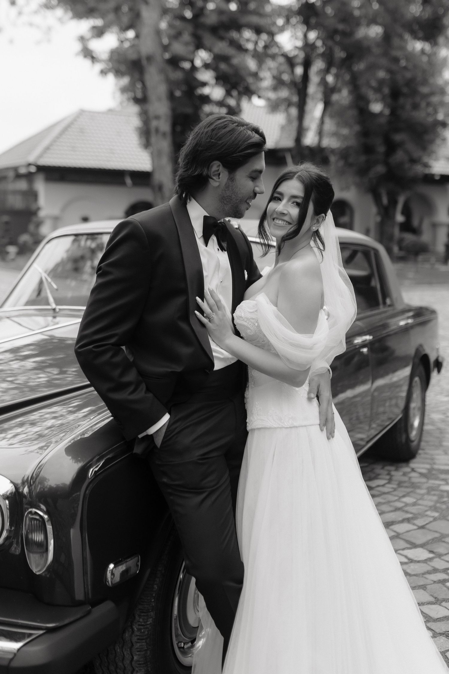 A romantic black and white wedding photo sequence showing tender moments between newlyweds beside a classic car.