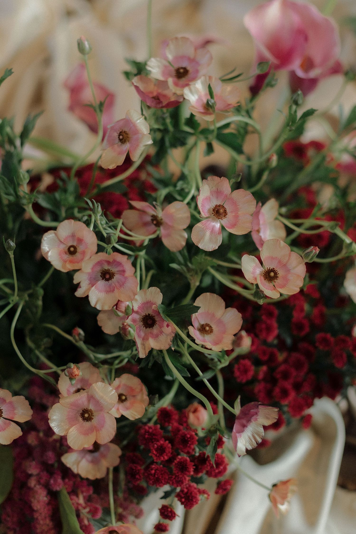 Pink anemone flowers with dark centers arranged with deep red blooms in a soft vintage-style floral arrangement.