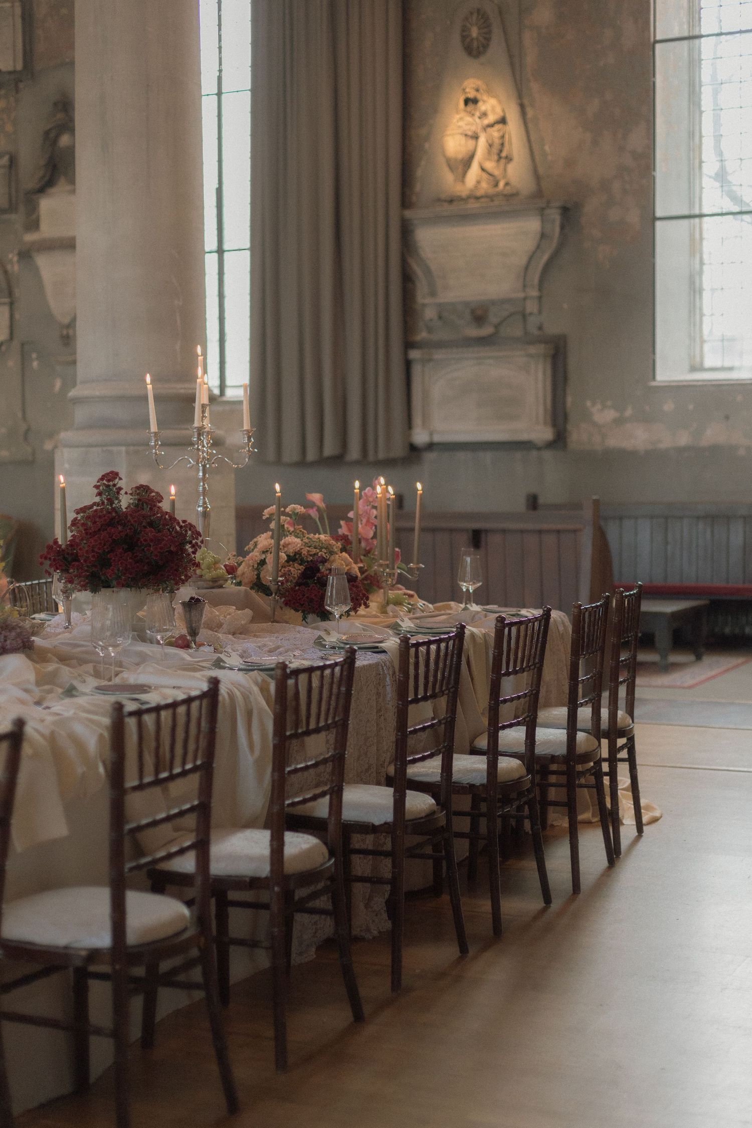 Elegant wedding reception tables with dark wood chairs and floral centerpieces line a grand hall with ornate walls and windows.