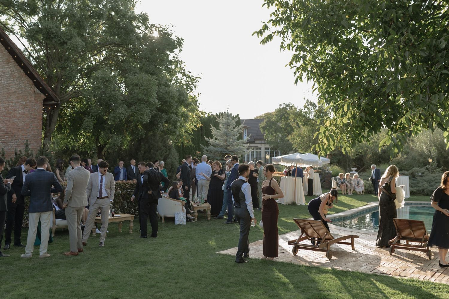 Guests mingle on a green lawn at an outdoor evening gathering with vintage cars parked nearby.