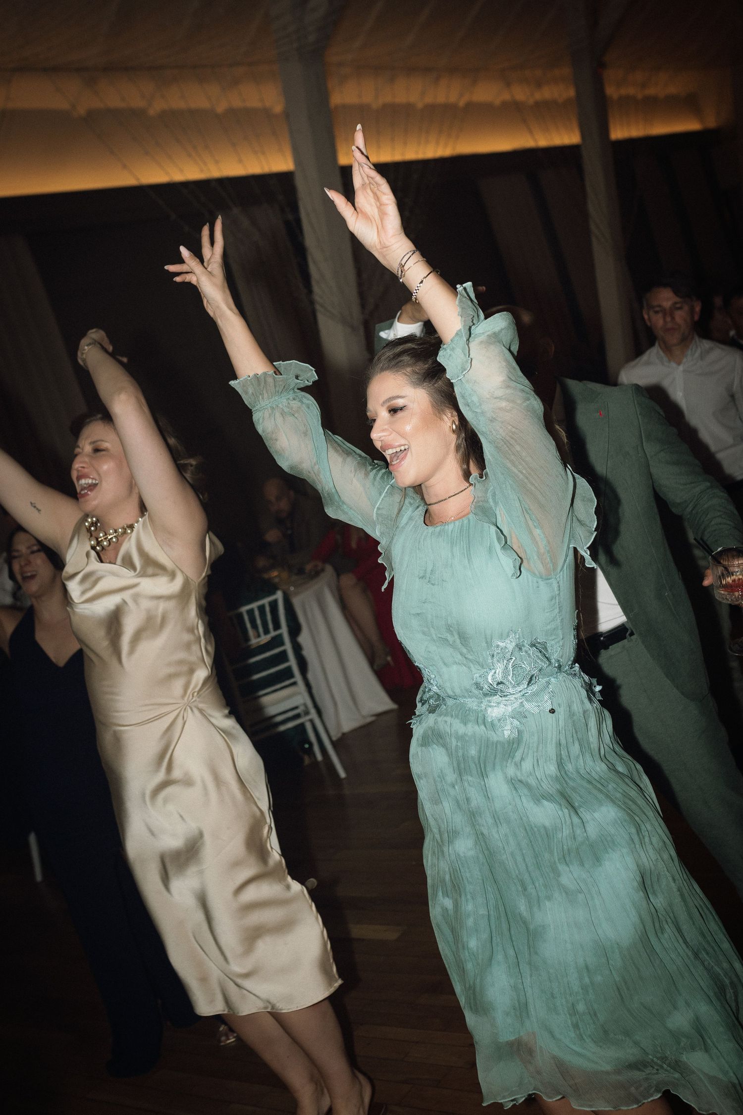 Two dancers in mint green and beige dresses move energetically on a dark dance floor during a celebration.