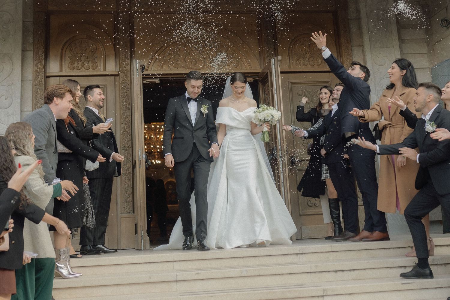 Wedding party celebrates with confetti and cheers as newlyweds exit historic church on stone steps.