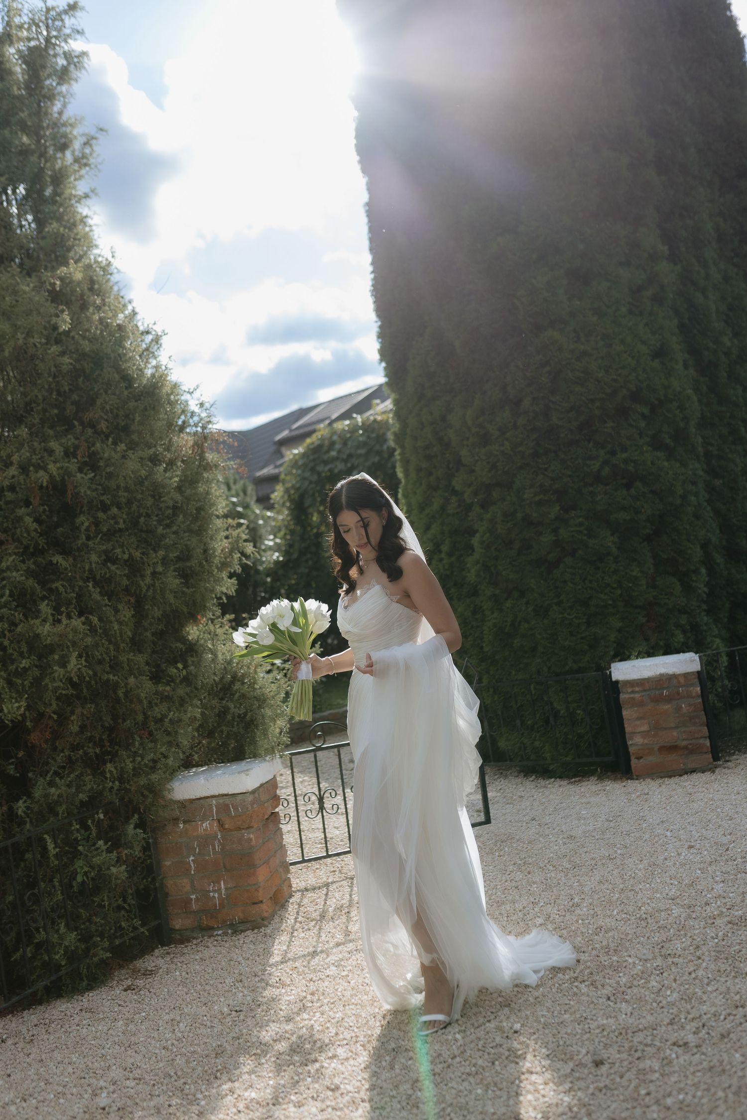A bride in a flowing white wedding dress walks along a gravel path bordered by tall hedges on a sunny day.