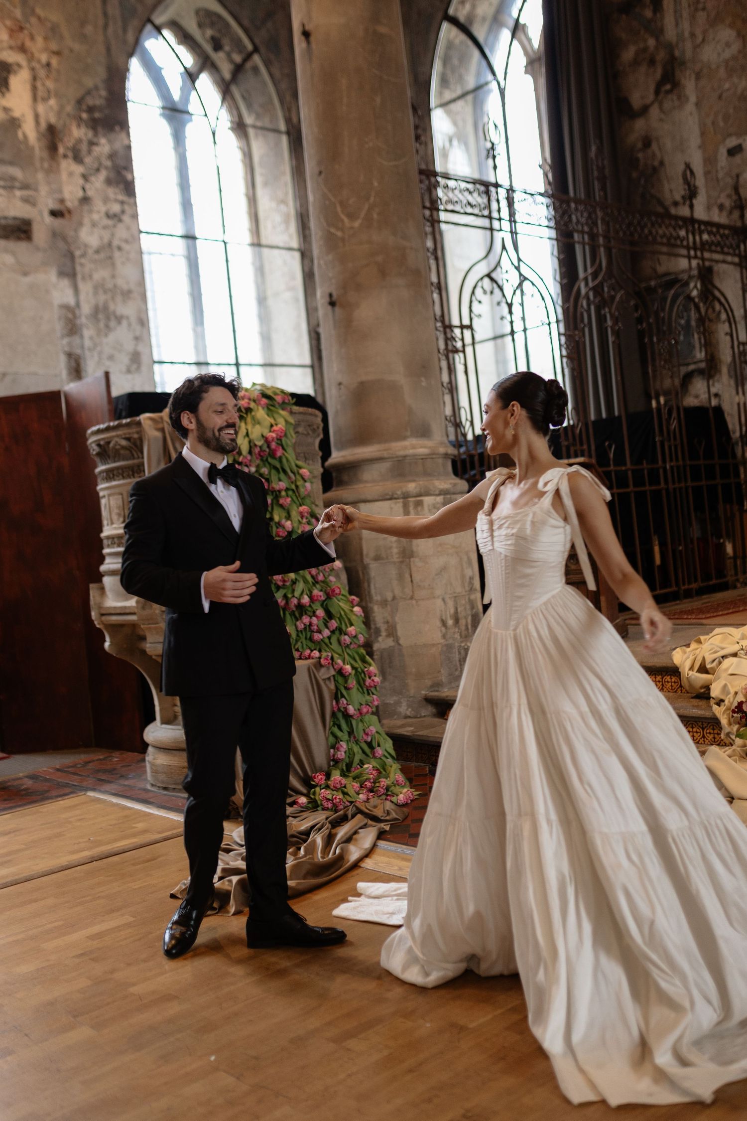 A couple in wedding attire dance together in a historic church with gothic windows and stone walls.