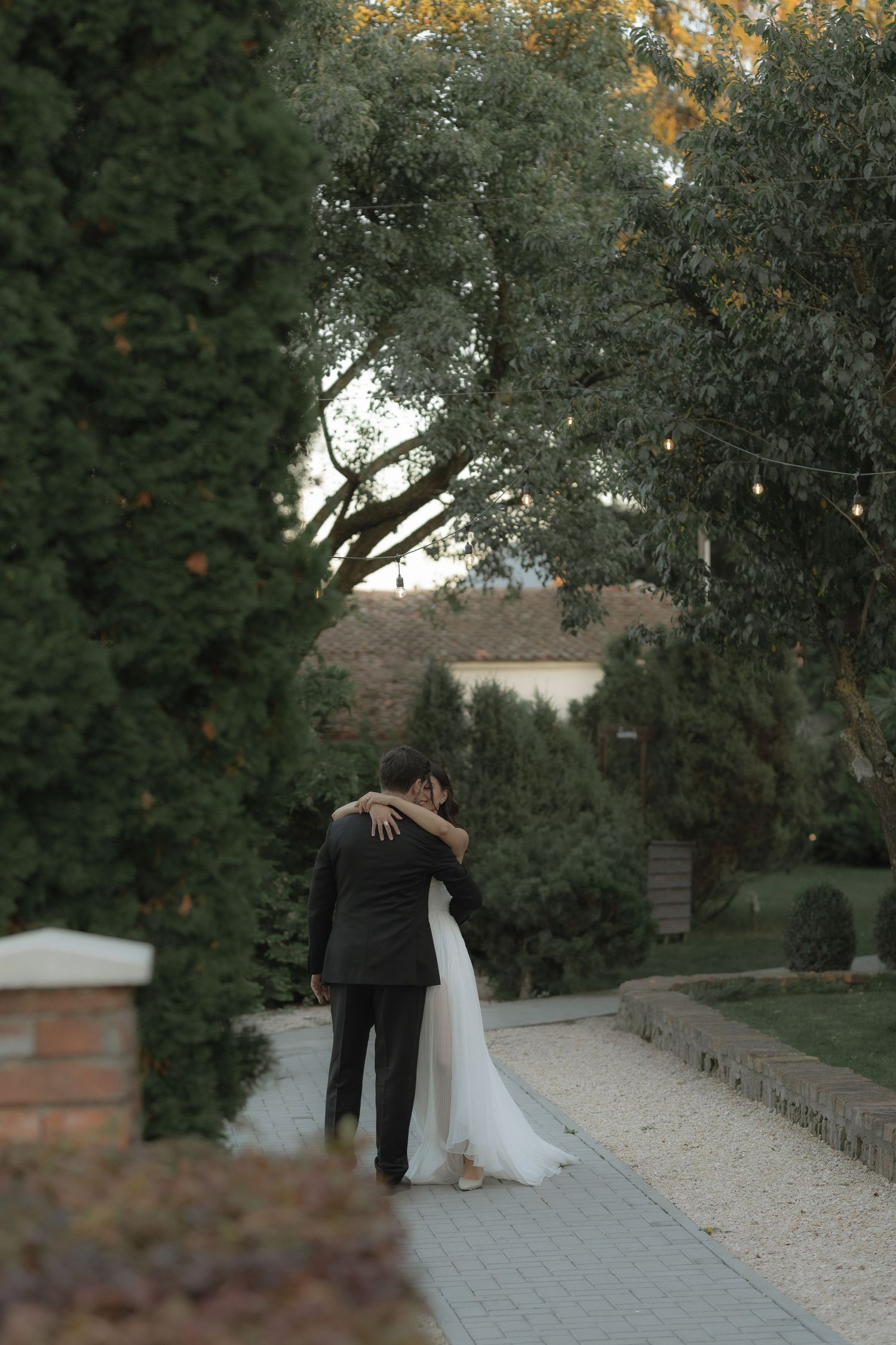 A romantic wedding couple walks down a tree-lined stone path at sunset in a garden setting.