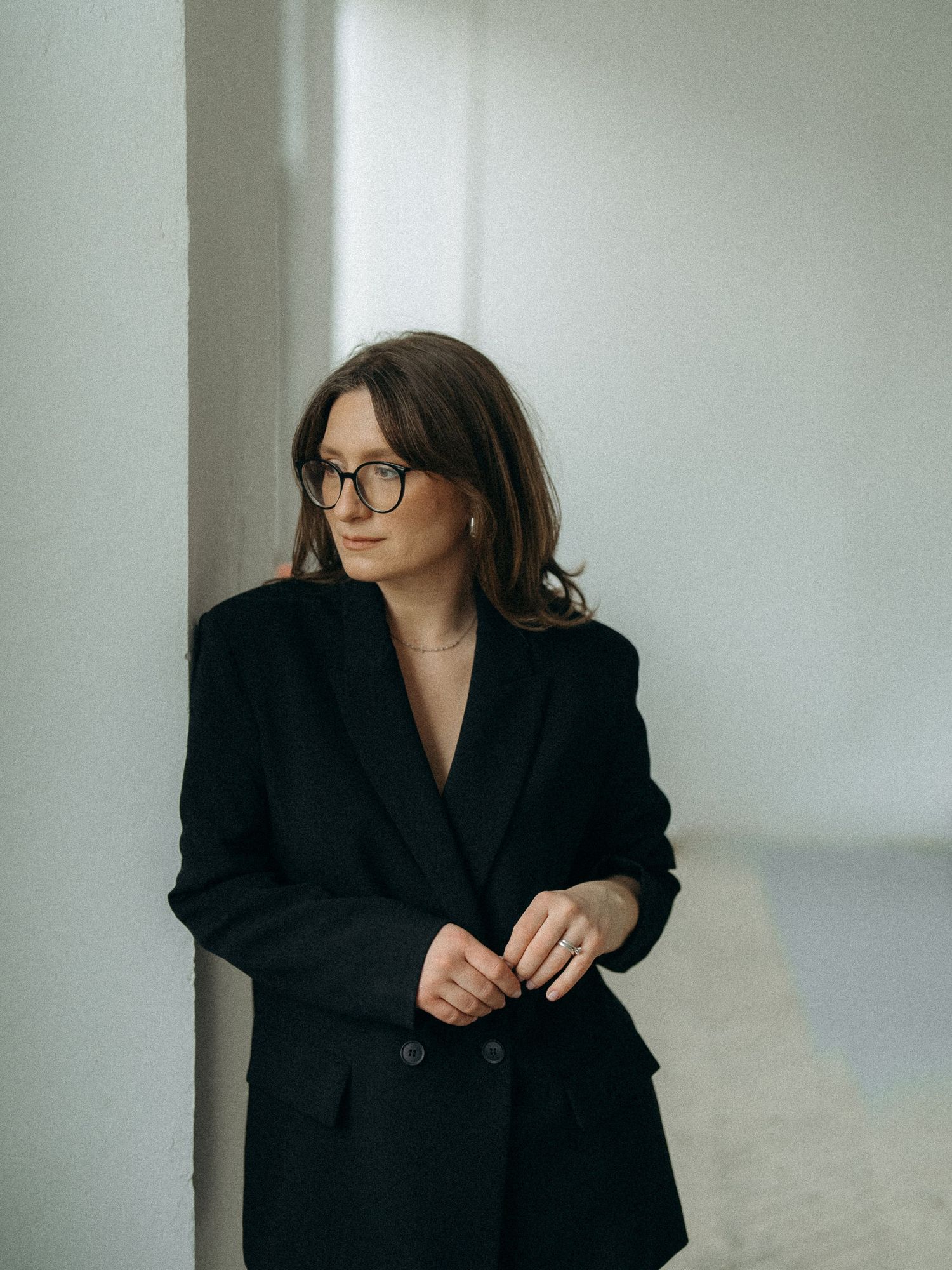 A person in a black blazer and glasses poses thoughtfully against a white wall in an artistic portrait series.