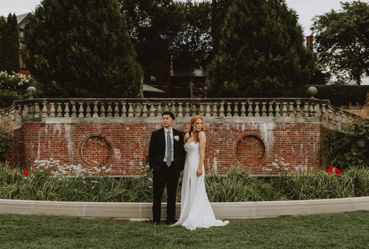 Bride and groom stand elegantly before a historic brick garden wall with blooming flowers and tall trees.