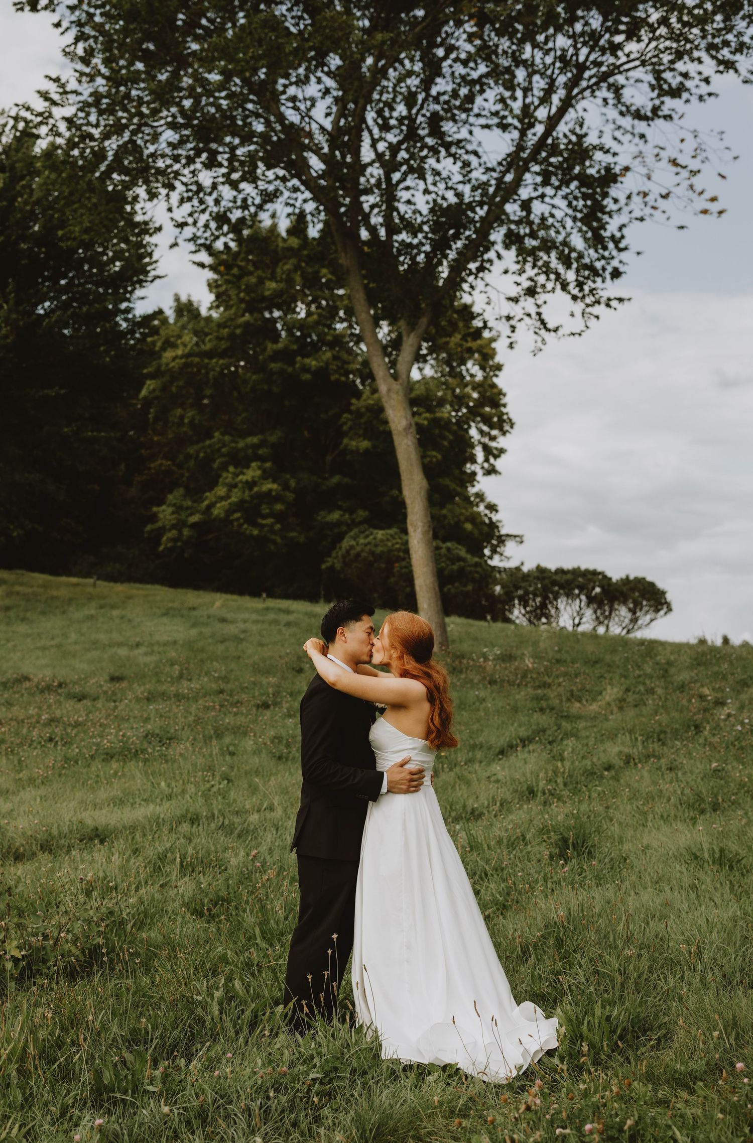 Bride and groom embrace romantically near a tall tree on a green hillside at golden hour.